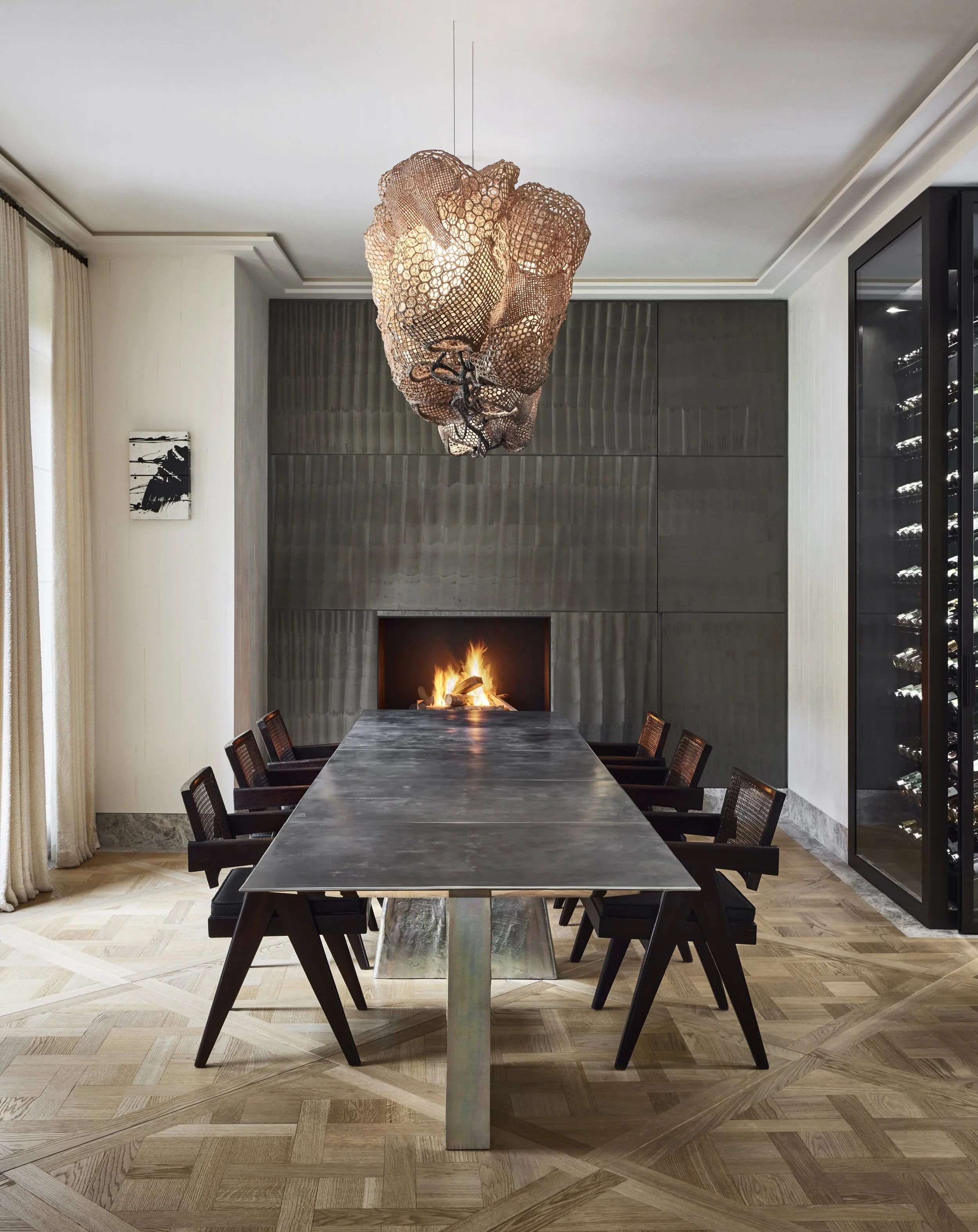 Salle de réunion avec une table longue, huit chaises noires, un mur en pierre, un feu de cheminée, un chandelier contemporain suspendu au plafond, et un mur vitré avec une cave à vin.