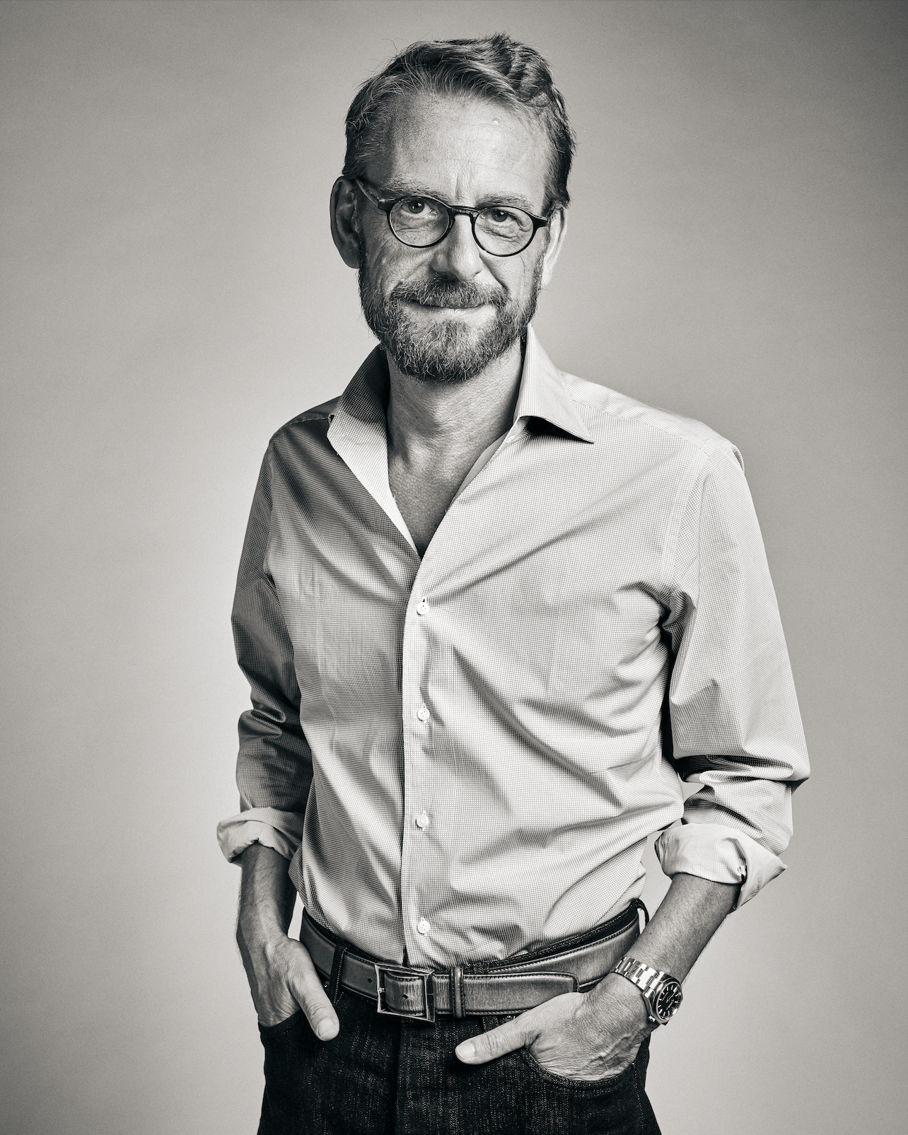 Portrait en noir et blanc d'un homme avec une barbe, des lunettes, portant une chemise à manches longues et un bracelet, posant avec les mains dans les poches.