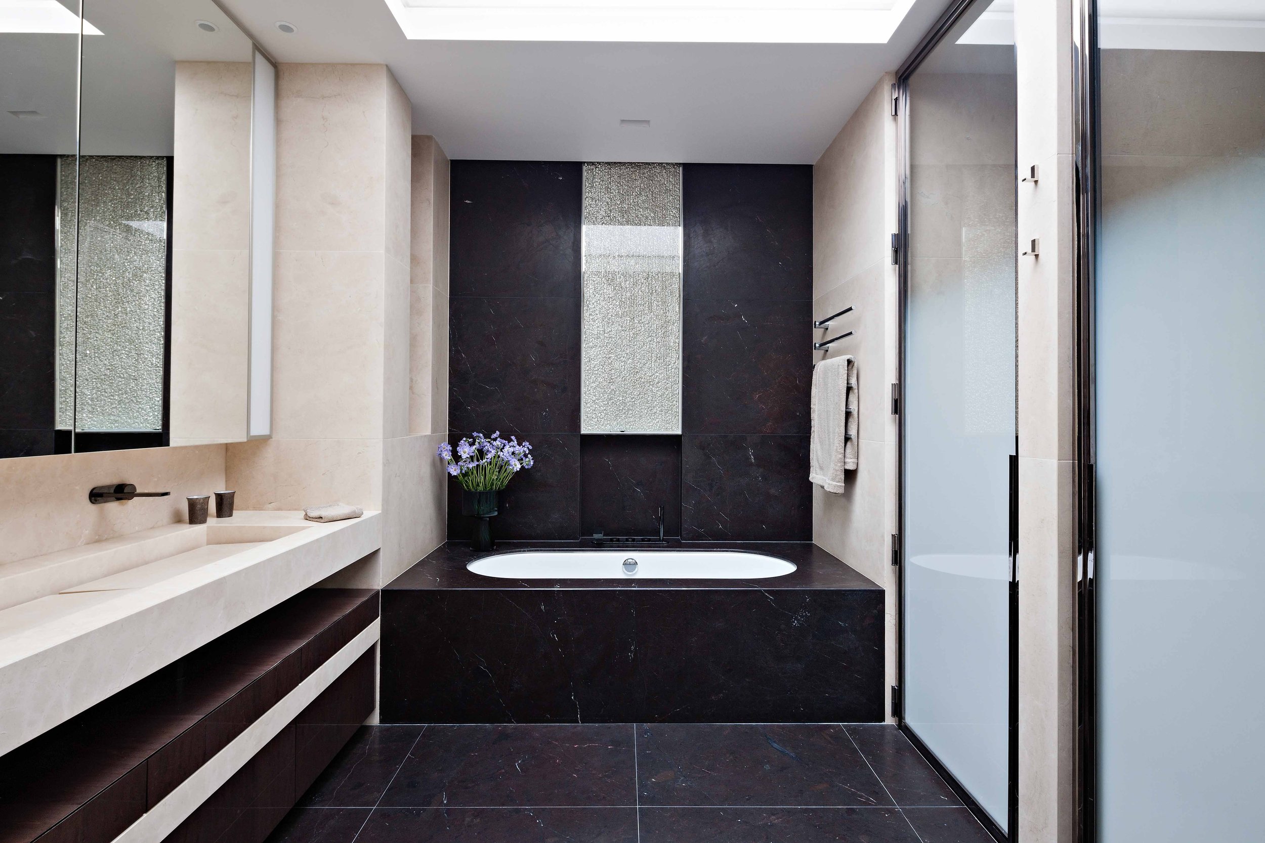 Salle de bain moderne avec baignoire encastrée en marbre noir, grand miroir, lavabo beige, et porte en verre dépoli.