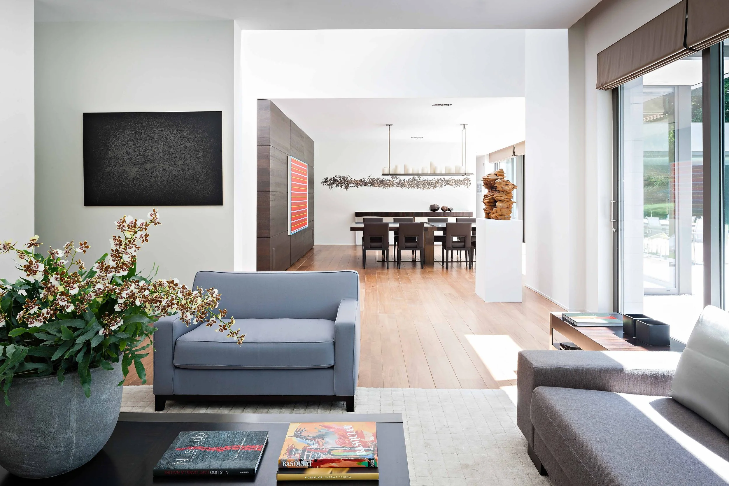 Salon spacieux avec canapés gris, table basse ornée de livres, vase avec des fleurs, et une salle à manger en arrière-plan avec une longue table et des chaises, éclairé par de grandes fenêtres laissant entrer la lumière naturelle.