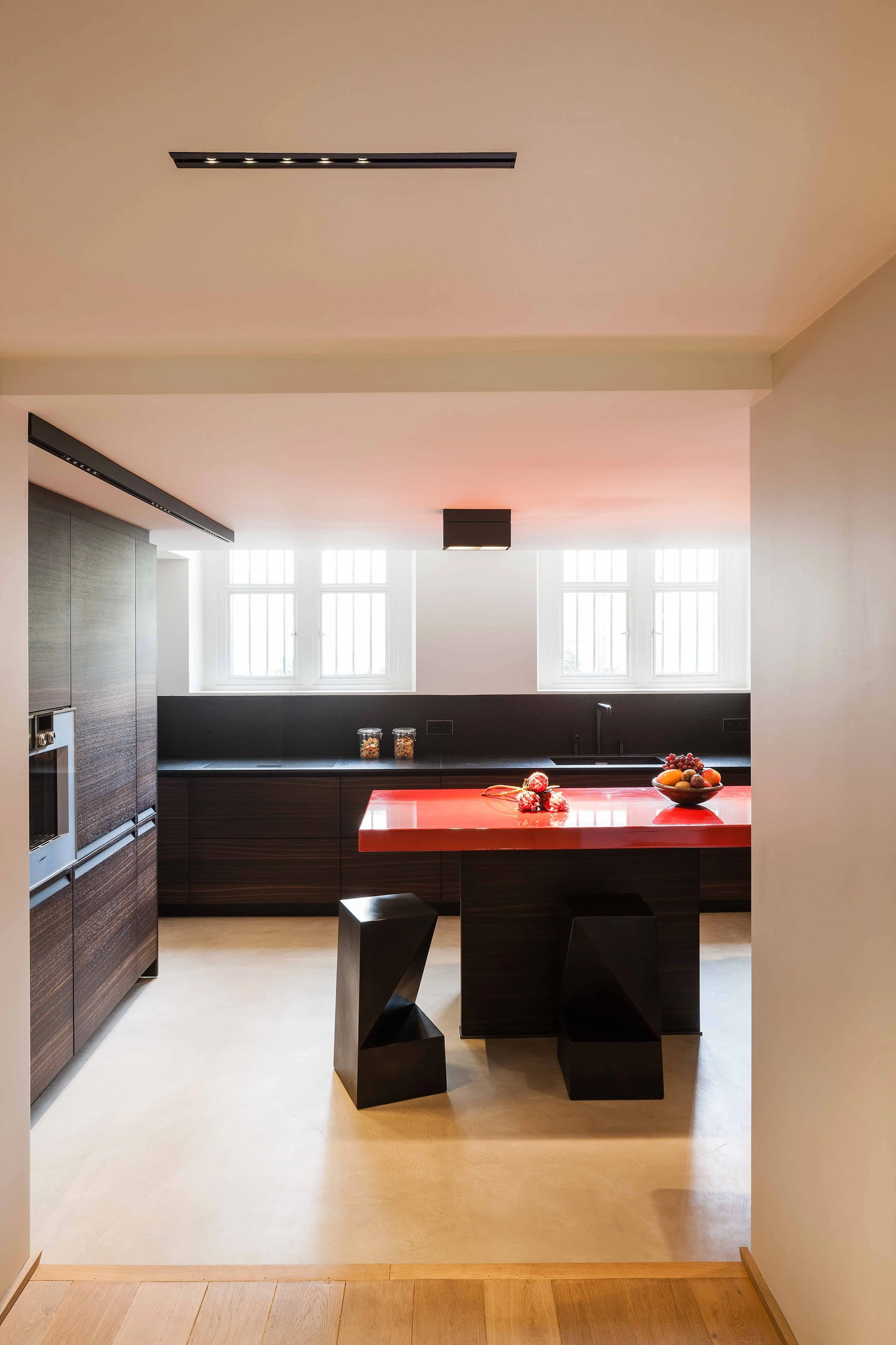 Cuisine moderne avec comptoir noir, fruits dans un bol, lumière naturelle, meubles en bois foncé, table rouge avec deux assises