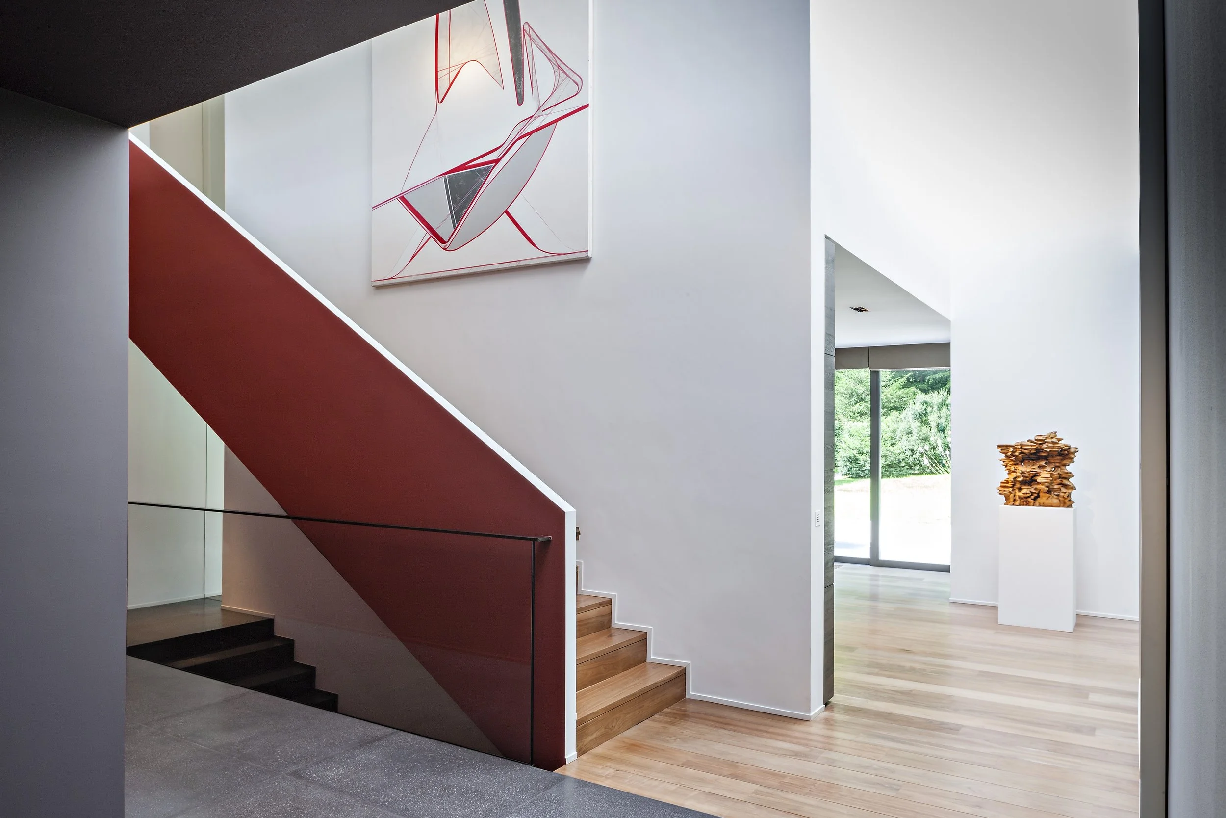 Intérieur moderne d'une maison avec escalier en bois, tableau abstrait accroché au mur, sculpture en bois sur un piédestal, grande fenêtre laissant entrer la lumière naturelle.