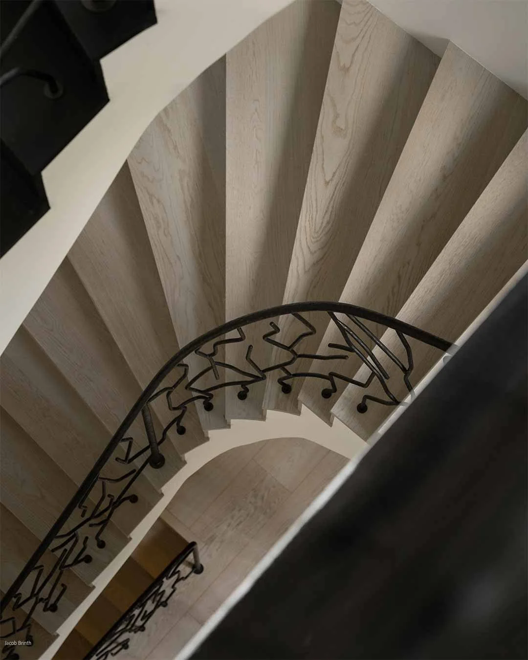 Vue du haut d'un escalier en colimaçon avec des marches en bois clair et une balustrade en métal noir.