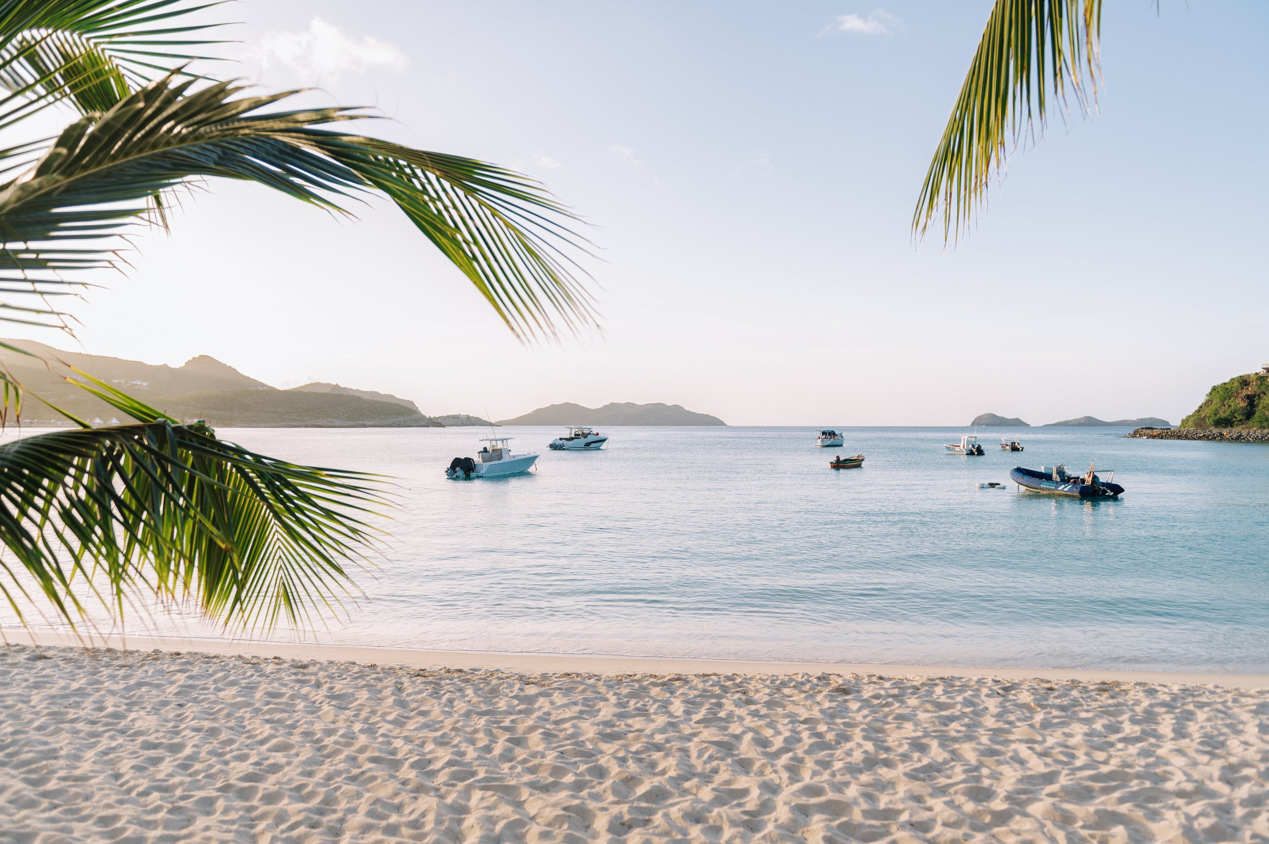 Plage avec sable, palmiers, et bateaux au large, en arrière-plan des montagnes et un ciel clair.