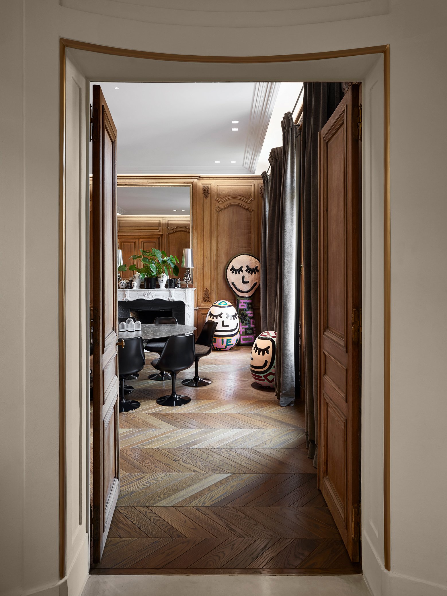 Une salle à manger élégante avec un parquet en bois clair, des rideaux gris foncé, un feu de cheminée blanc avec un vase vert dessus, et des sculptures modernes barbijolleurs avec des visages souriants et des motifs colorés.