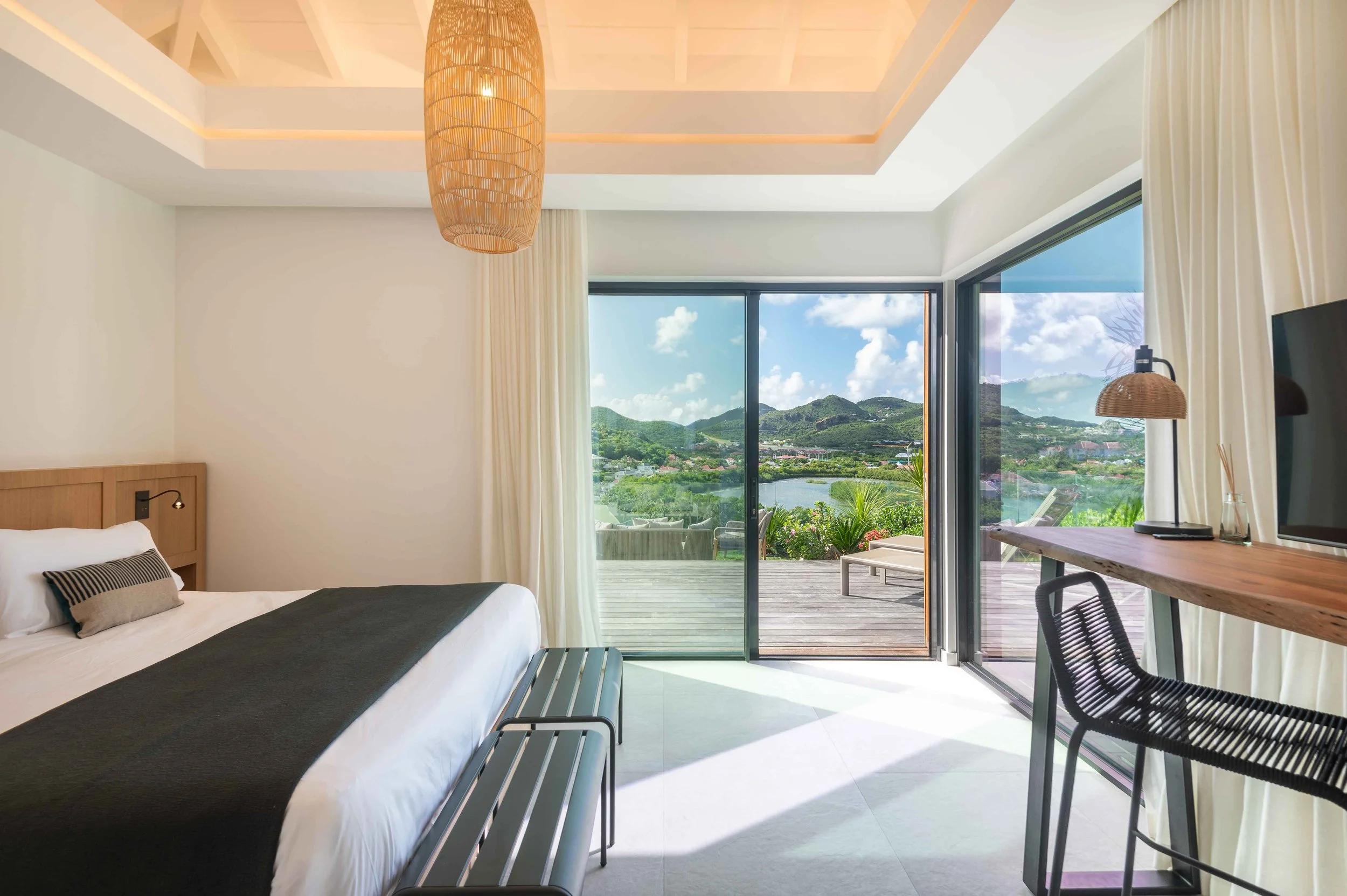 Chambre moderne avec lit blanc, vue sur paysage verdoyant, mobilier en bois naturel, lampes en rotin et balcon extérieur.