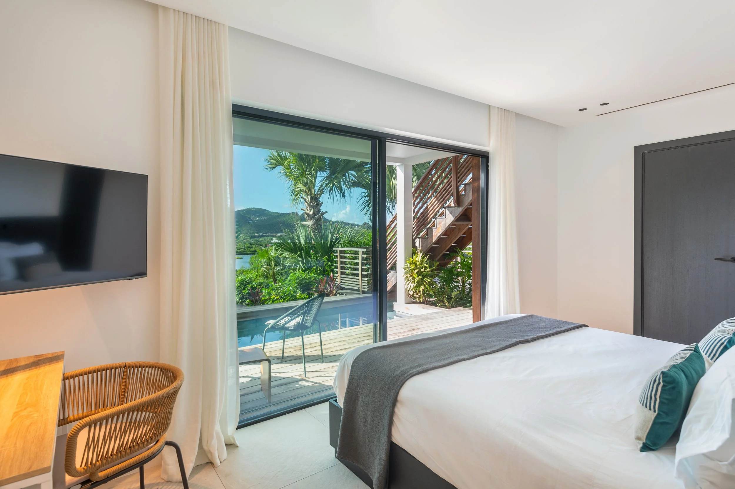 Chambre moderne avec lit, vue sur terrasse extérieure avec plantes, chaise et vue sur nature, porte-fenêtre avec rideaux blancs.