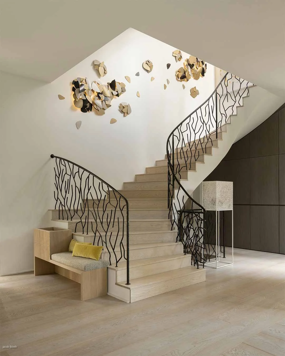 Escalier en bois avec rampe en métal noir, décoration murale abstraite en forme de pétales ou feuilles, lampe moderne avec abajur en pierre, coin avec banc en bois clair et coussin jaune, sol en bois clair, murs blanc, côté droit avec panneau en bois