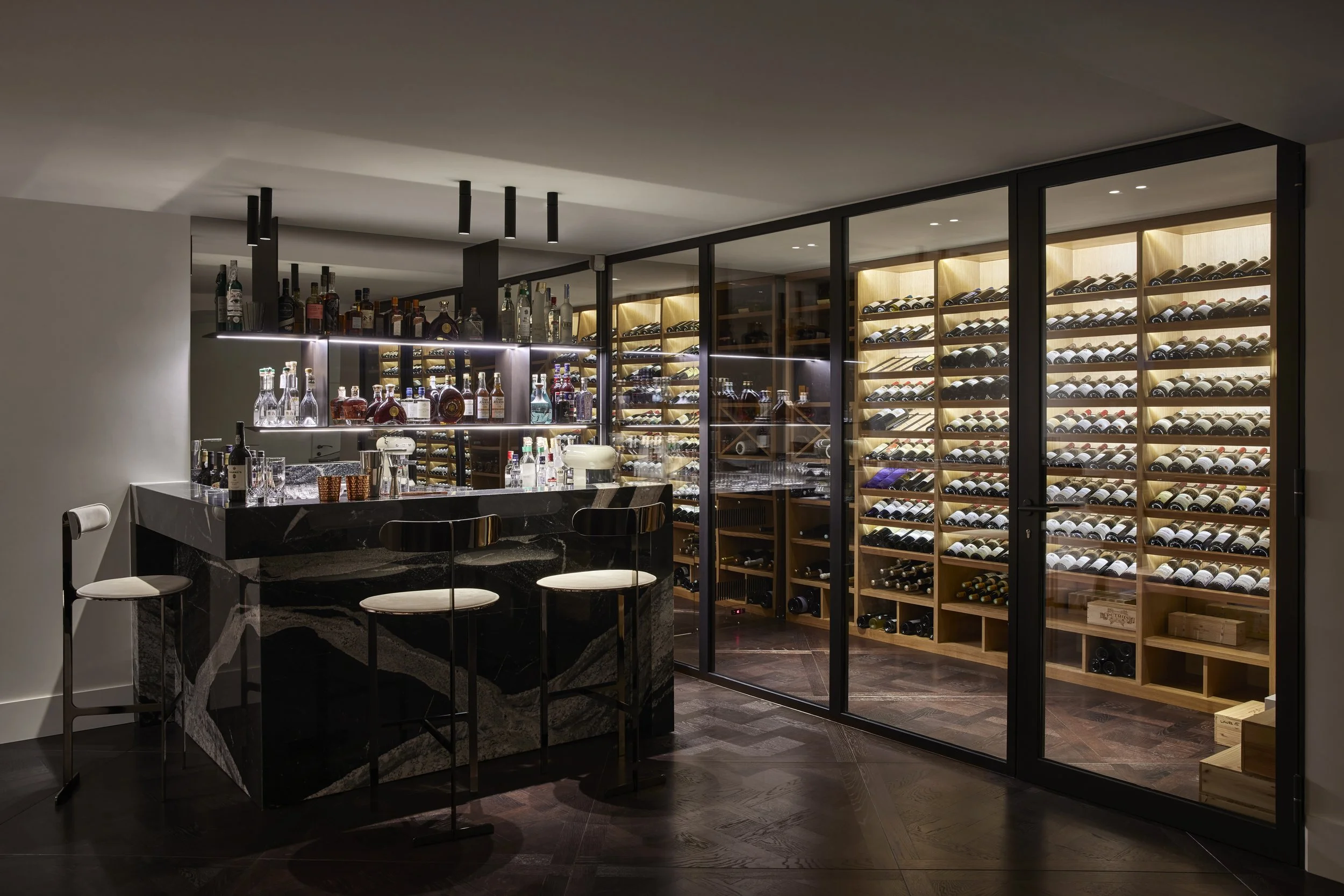 Un bar avec un comptoir en pierre noire et une armoire pour vin en bois avec porte en verre. Le bar est équipé de diverses bouteilles d'alcool et de glaçons, avec trois tabourets blancs autour du comptoir.