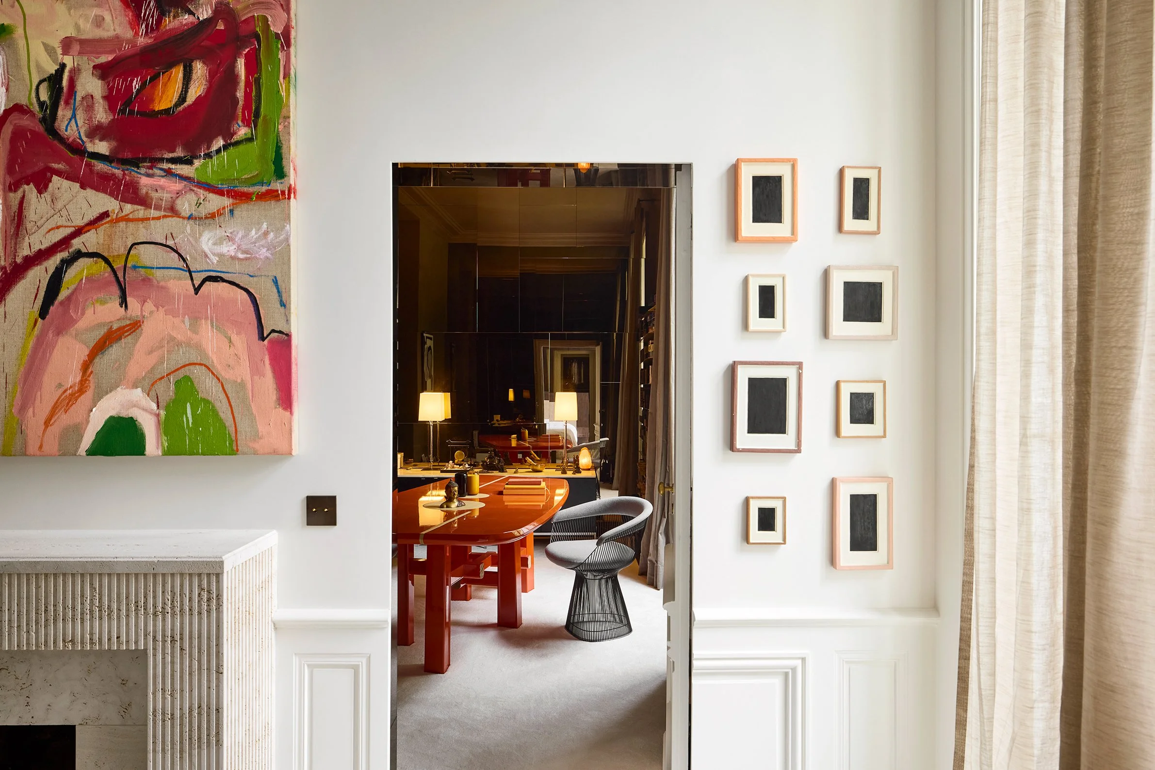 Vue intérieure d'une pièce avec mur blanc, œuvres d'art modernes et une ouverture vers une salle avec un grand bureau rouge et une chaise noire.