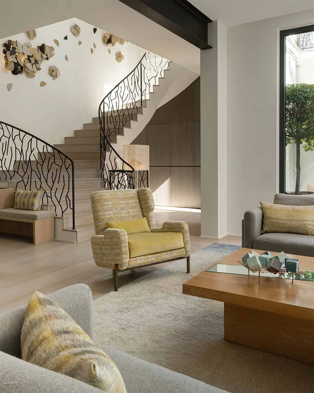 Salon moderne avec fauteuils, canapé et table en bois, escalier en spirale avec rampe décorative en métal noir, mur avec décoration murale en forme de fleurs, grande fenêtre laissant entrer la lumière naturelle, vue sur un arbre à l'extérieur.