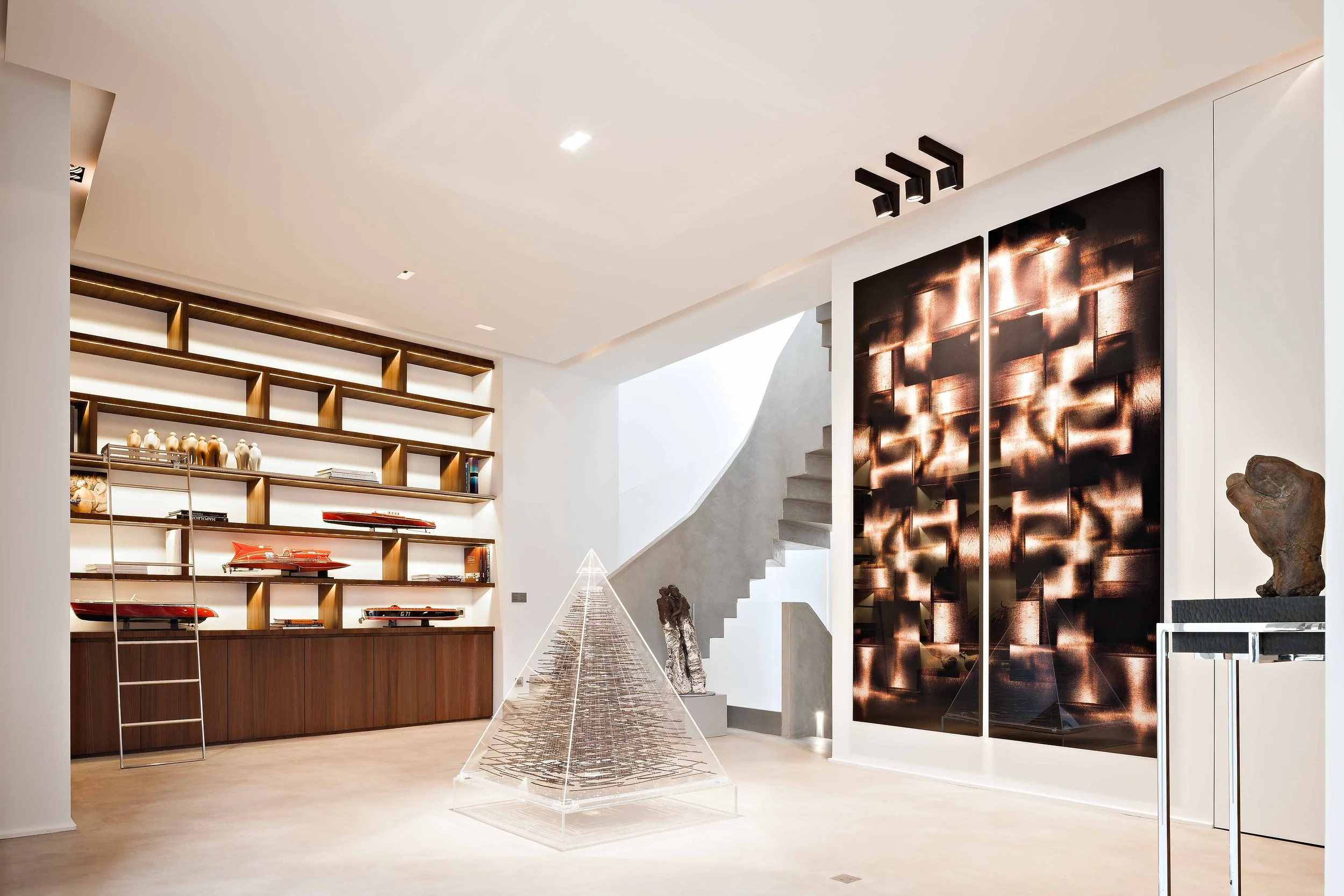 Intérieur moderne d'une maison avec étagères arrières en bois, sculpture en forme de pyramide en acrylique, porte murale avec motif lumineux, et escaliers en béton.