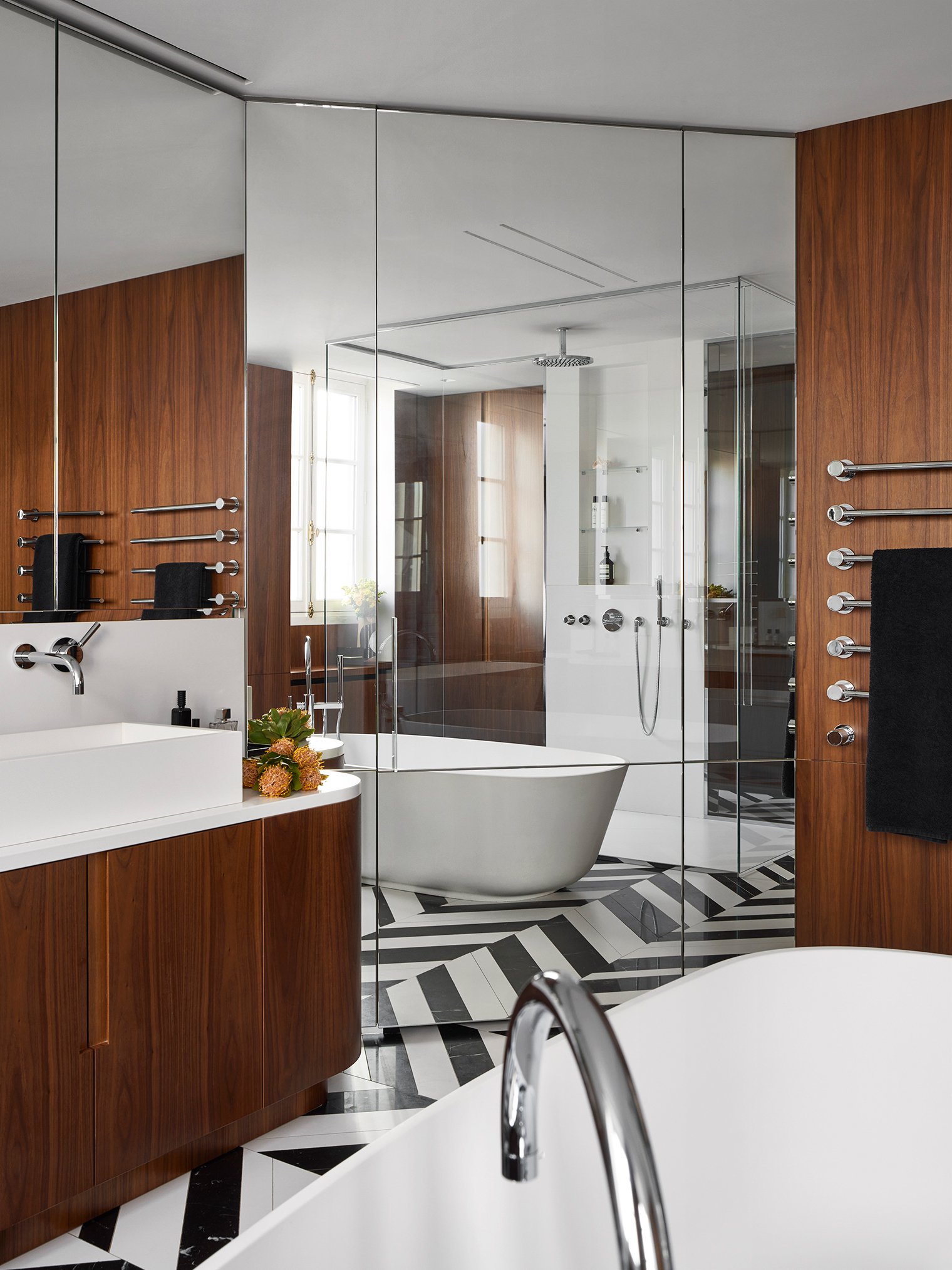 Salle de bain moderne avec baignoire blanche, murs en bois, sol à motif géométrique noir et blanc, douche séparée avec paroi en verre, miroir et accessoires en chrome, fenêtres avec rideaux, et touches décoratives telles que des fleurs sur le lavabo.