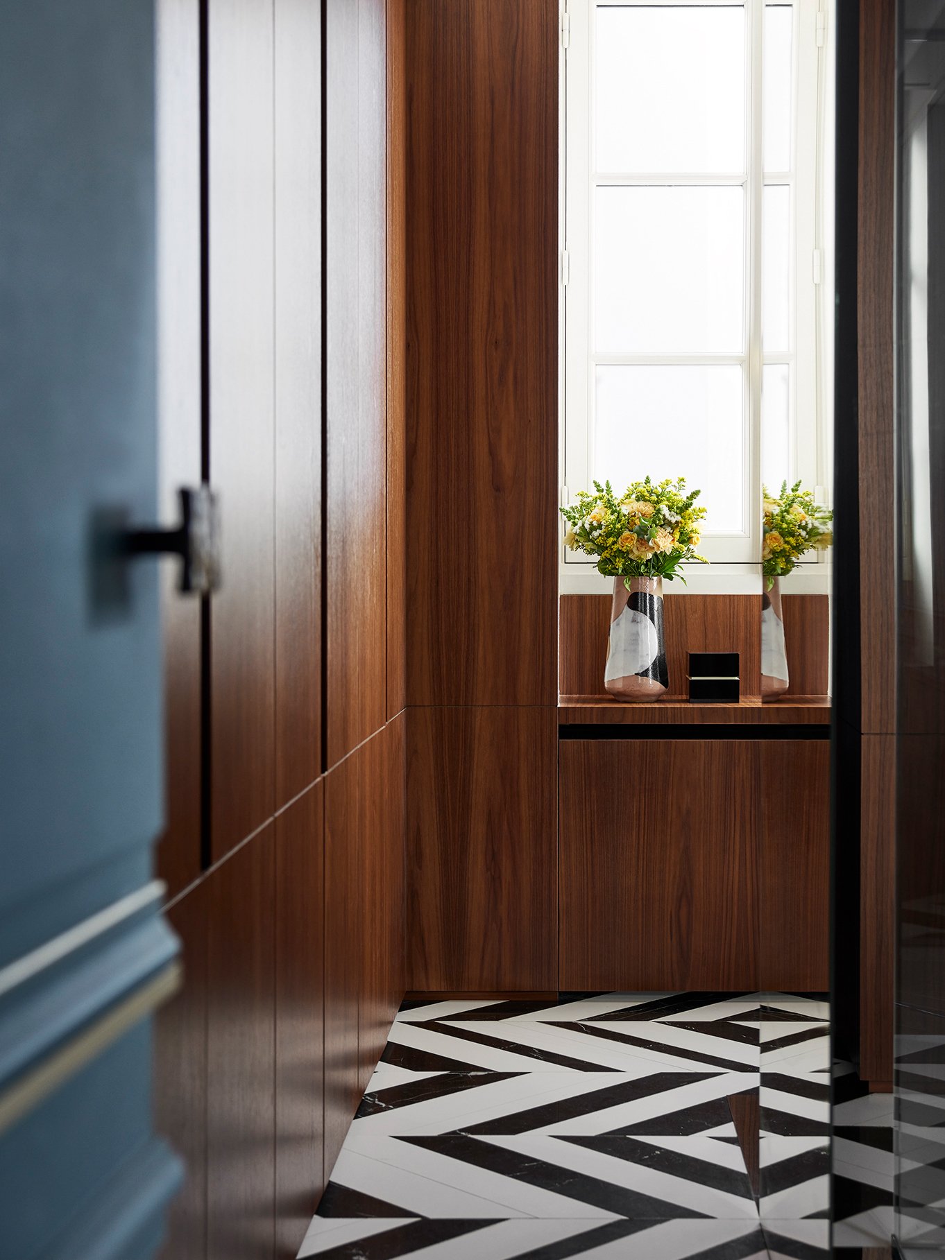 Intérieur d'une pièce avec des murs en bois clair et un sol en mosaïque noir et blanc, avec des vases de fleurs jaunes et blanches devant une grande fenêtre