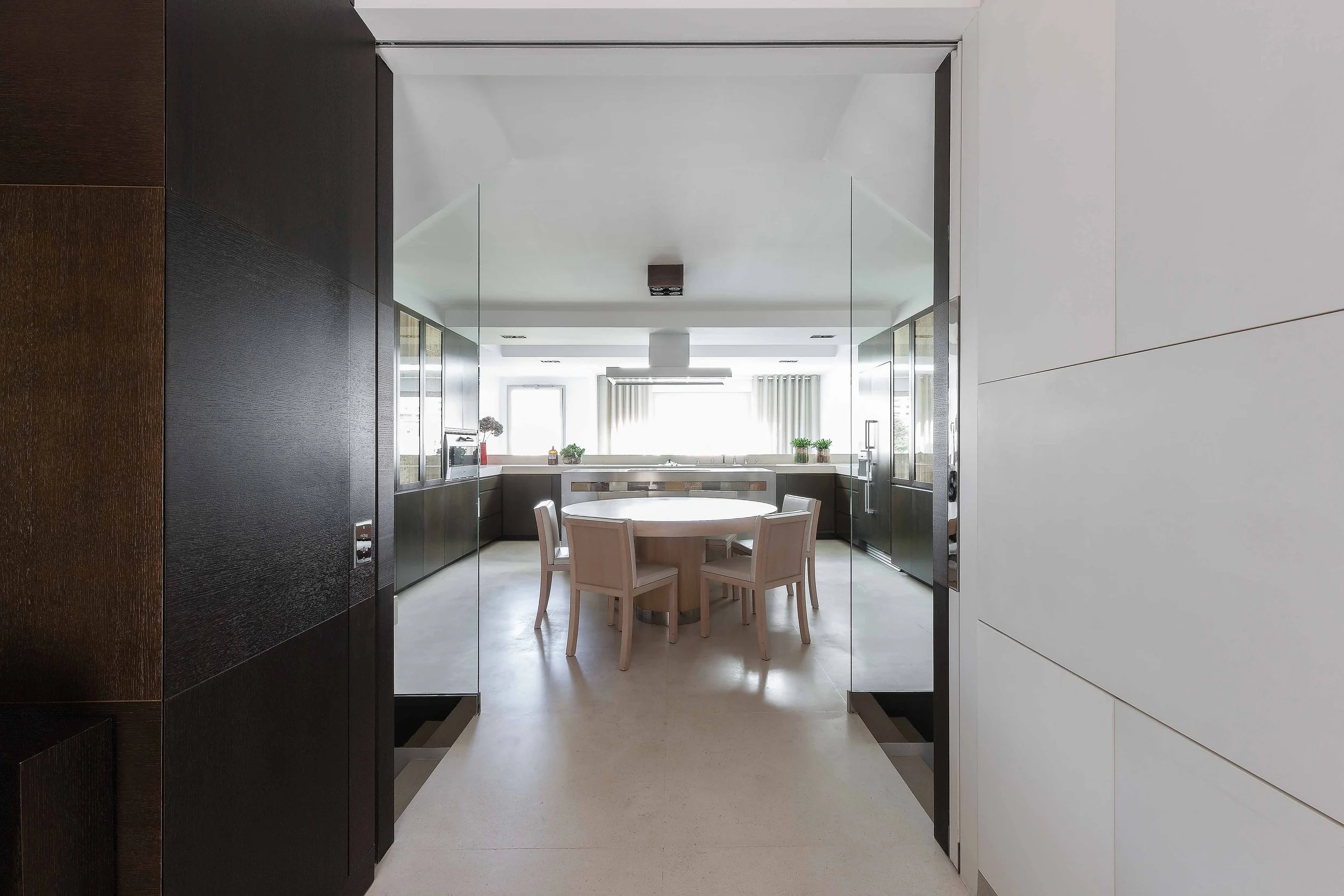 Vue d'une cuisine moderne vue à travers une porte en verre, avec un meuble de cuisine noir, une table ronde en bois clair et des chaises en bois clair, des plantes et des fenêtres lumineuses.