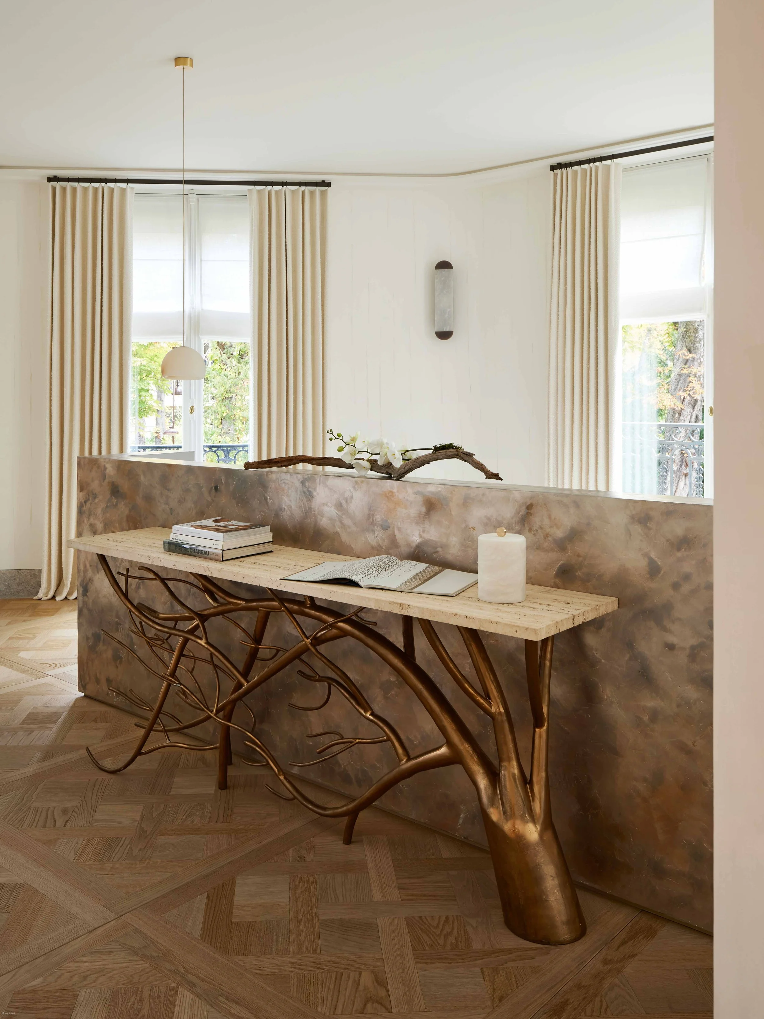 Intérieur d'un salon avec deux fenêtres, rideaux beige, table en bois avec base en forme de branches d'arbre en cuivre, décorations comme livres, bougie et branche avec fleurs, mur blanc