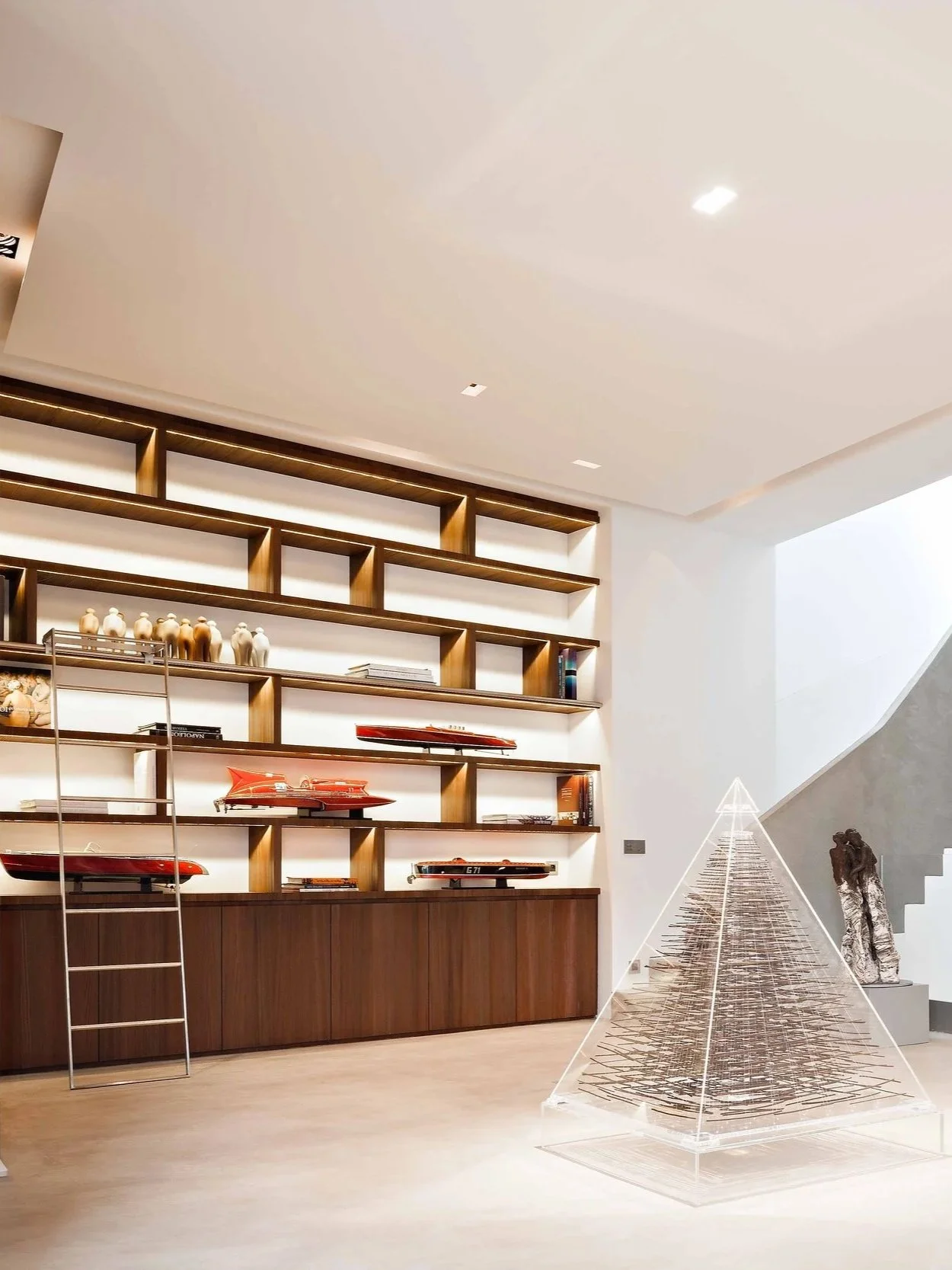 Une pièce moderne avec une grande bibliothèque en bois contenant des maquettes de navires, des livres et quelques figurines, ainsi qu'une sculpture en pierre et un arbre de Noël en matériaux transparents et métalliques.