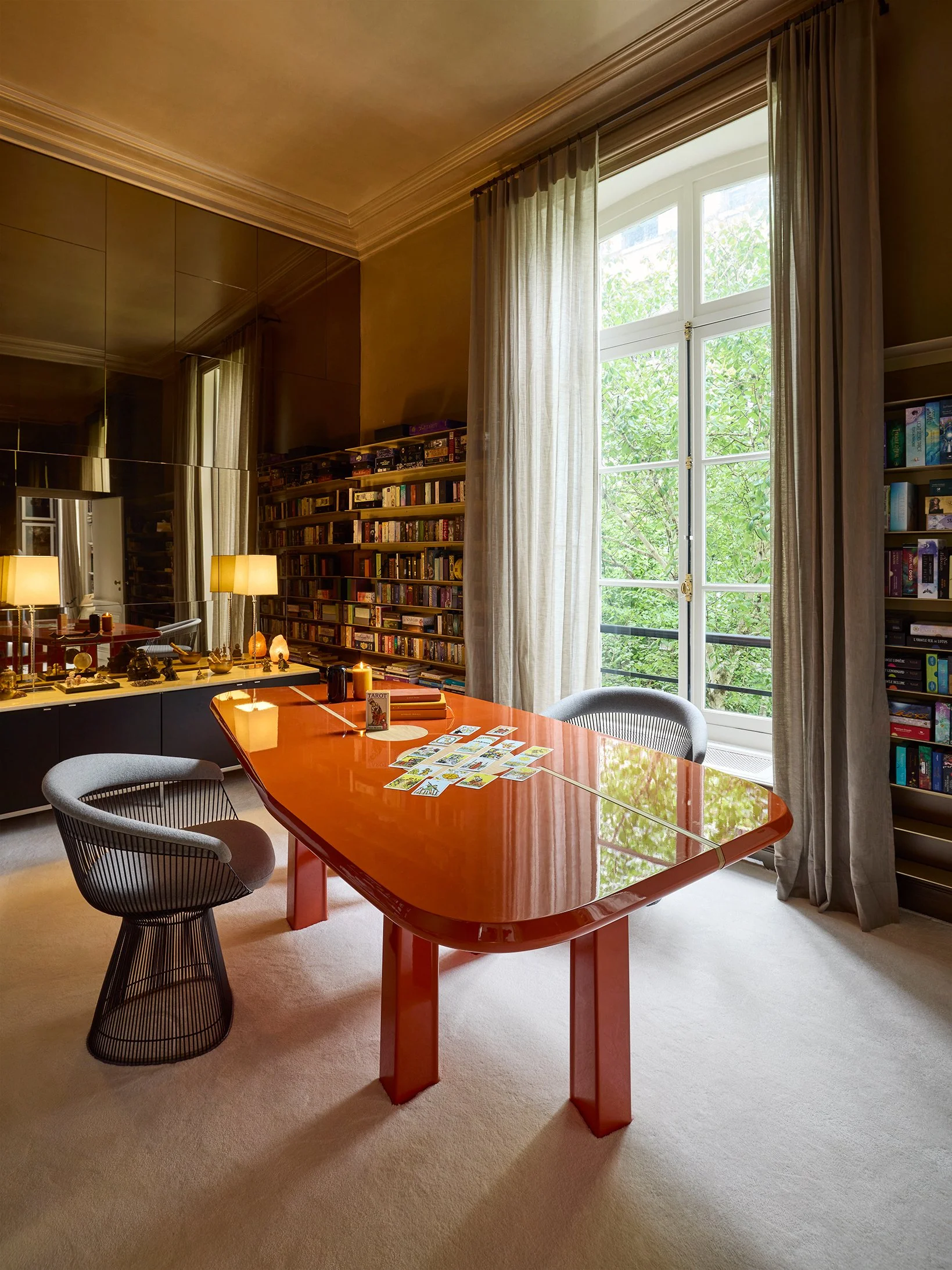Une pièce avec une grande fenêtre laissant entrer beaucoup de lumière naturelle, une table orange avec des cartes de tarot dessus, deux chaises, et des étagères remplies de livres.