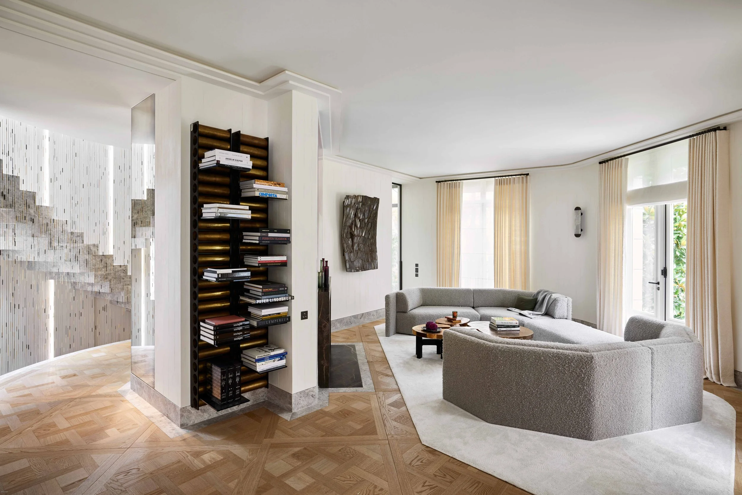 Salon moderne avec canapé gris, fenêtres avec rideaux beige, bibliothèque décorative noire et bronze, et tableau abstrait sur le mur.