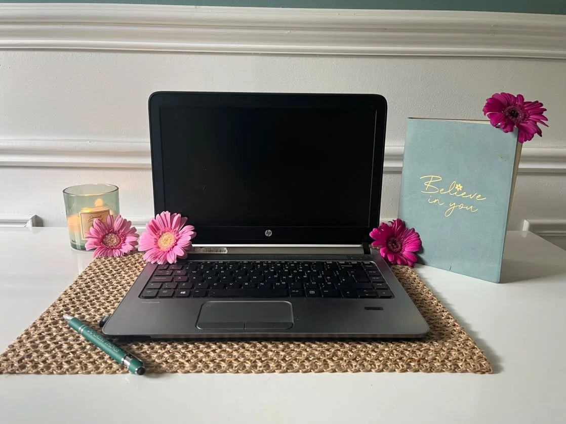 A laptop on a woven placemat with pink flowers, a candle, a teal pen, and a light blue sign that reads 'Believe in you' arranged on a white surface.
