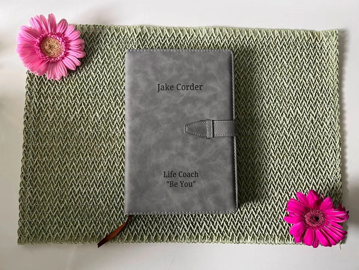 A gray leather planner with the name Jake Corder and the phrase 'Life Coach Be You' on the cover, placed on a green woven mat, with pink gerbera daisies on the top left and bottom right corners.