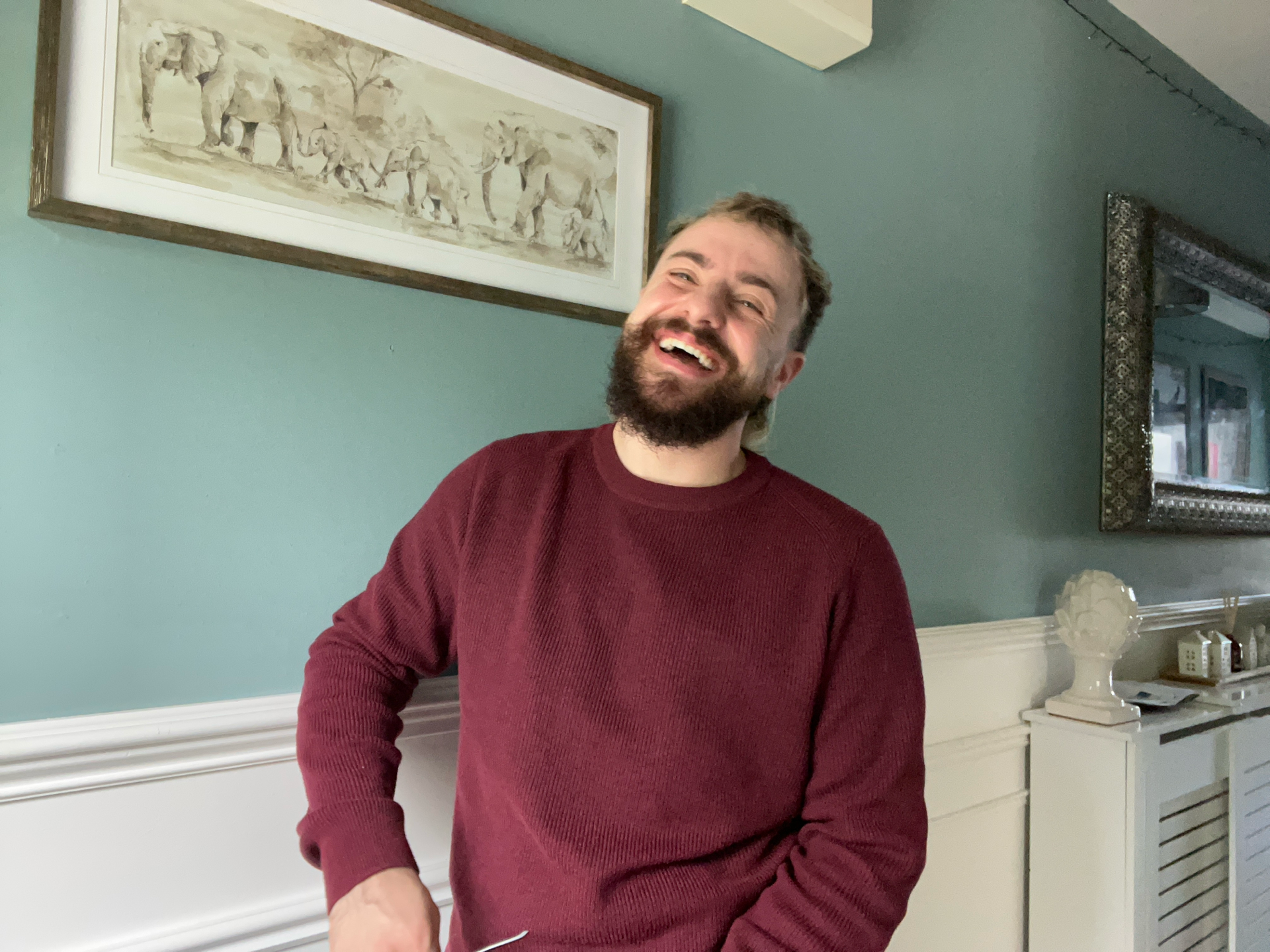 A man with a beard and short hair, wearing a maroon sweater, is smiling and looking directly at the camera. He is standing indoors against a teal wall, with a long framed artwork of elephants hanging behind him, and a mirror to his right. There is a white sideboard below the mirror with decorative items and an electronic device on it.