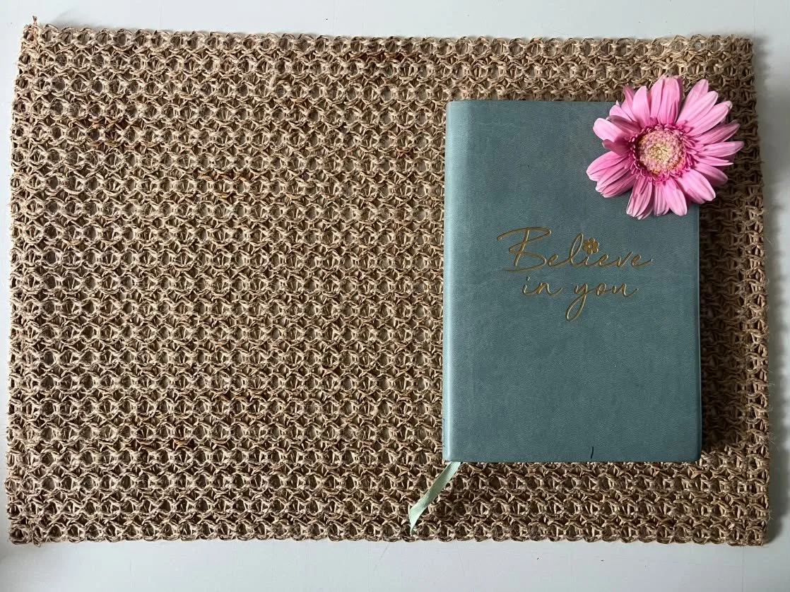A teal hardcover journal with gold lettering reading "Believe in you" is placed on a woven brown mat. A pink flower with a yellow center rests on the top right corner of the journal.