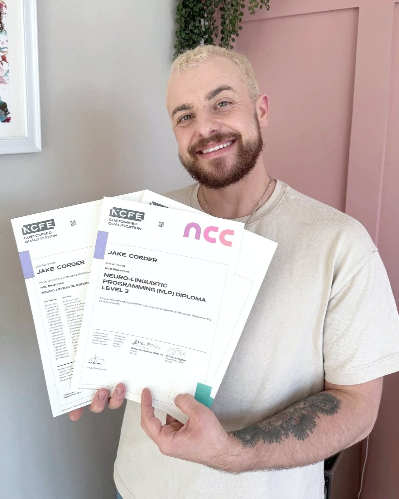 Jake is smiling holding multiple certification diplomas for Neuro-Linguistic Programming (NLP) Level 3, standing indoors with pink and gray walls, a framed picture, and some greenery in the background.