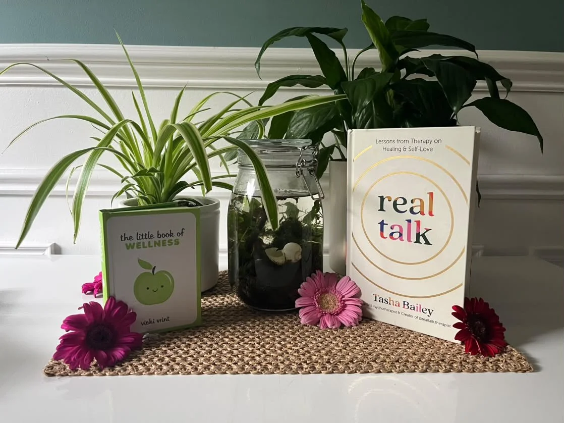 Two potted green plants, a glass jar with dried plants inside, a book titled 'real talk' by Tasha Bailey, and pink and red flowers on a woven mat on a white shelf.