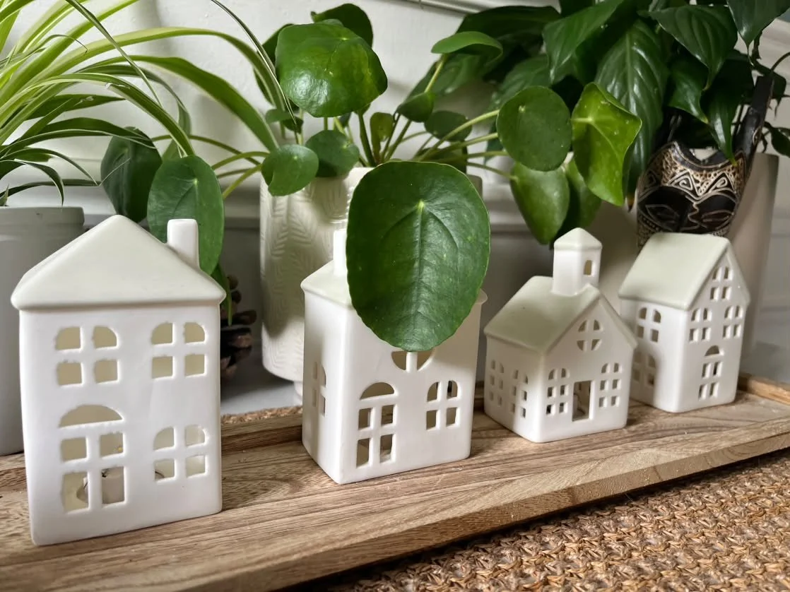 Decorative white ceramic houses with potted green plants in the background on a wooden surface.