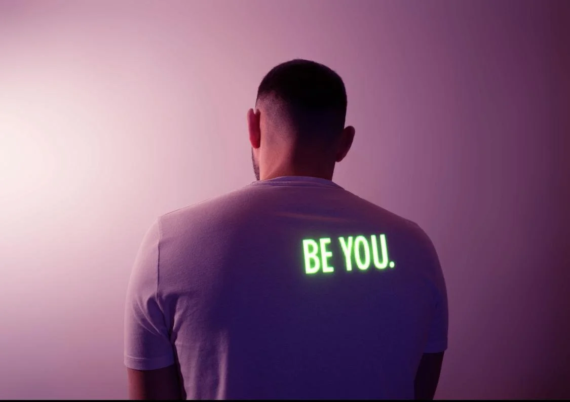 Back view of a man with short dark hair wearing a white t-shirt that glows with the words "BE YOU." on his back, against a pink and purple gradient background.