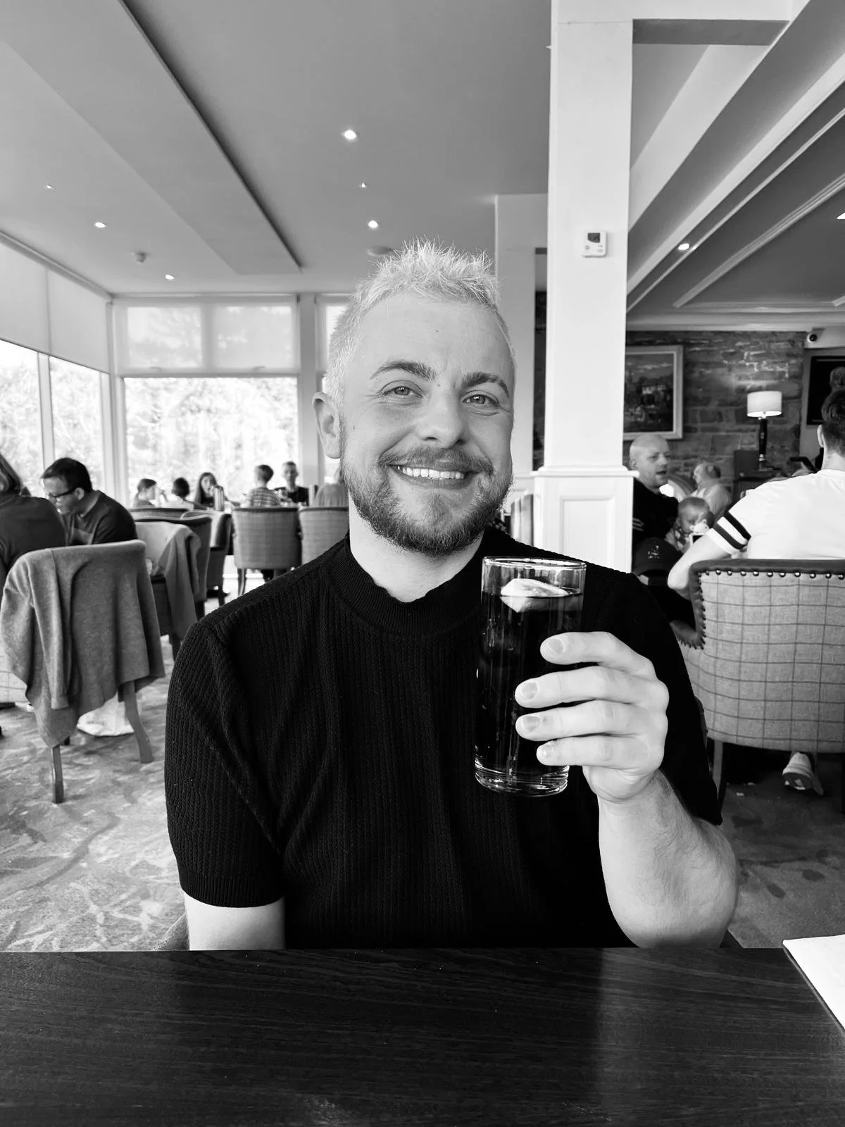 Jake smiling holding a glass of dark soda with ice, seated in a restaurant with people in the background.