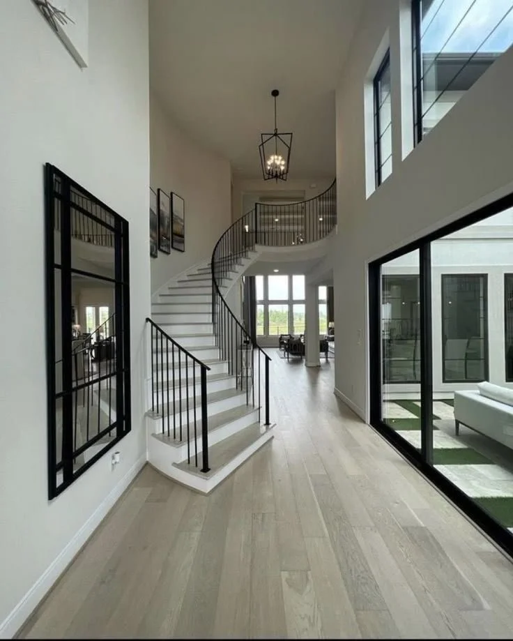 A modern, two-story open-concept home interior featuring a curved staircase with black railings, large windows, and minimalistic decor.