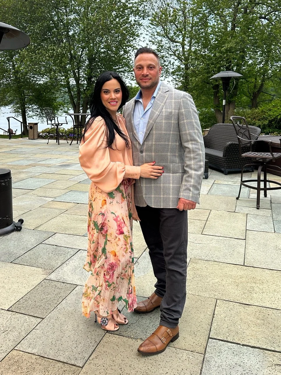 A couple standing outdoors on a stone patio, with trees and outdoor furniture in the background, smiling and posing for the photo.