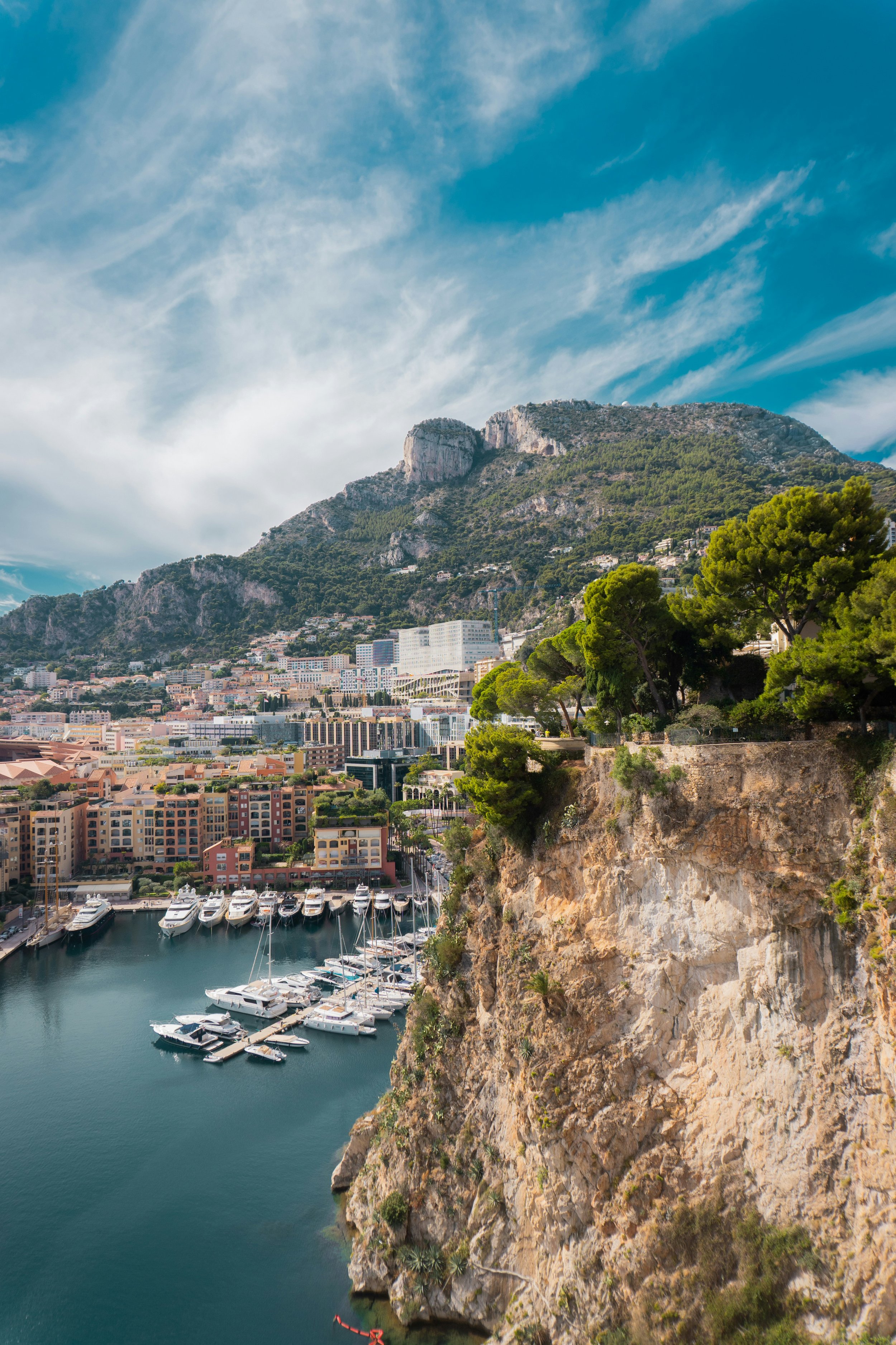 Docked yachts and boats in a marina next to multi-story buildings, with a hillside covered in trees and houses in the background under a partly cloudy sky.