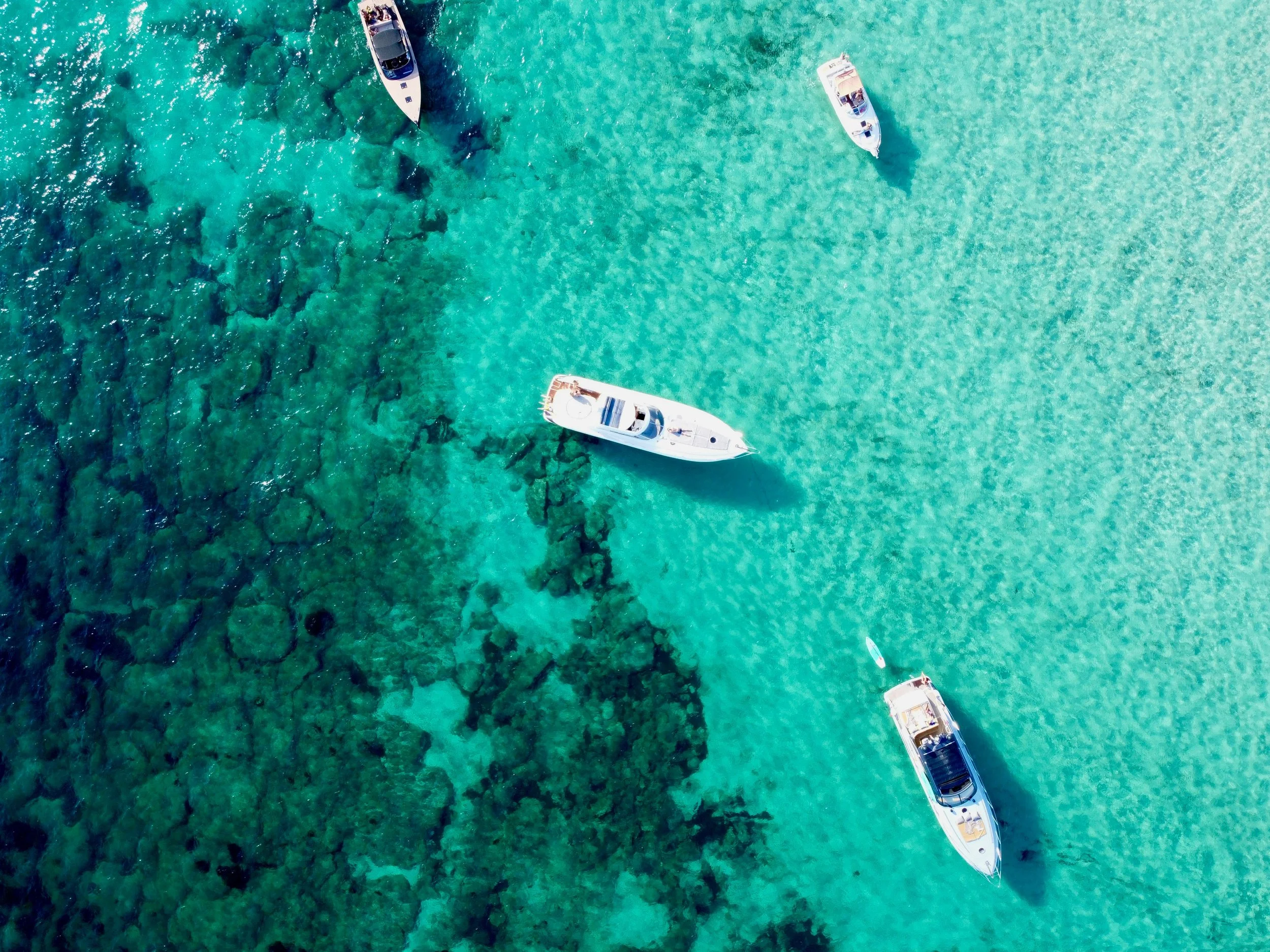Overhead view of several boats anchored in clear turquoise water near a coral reef.