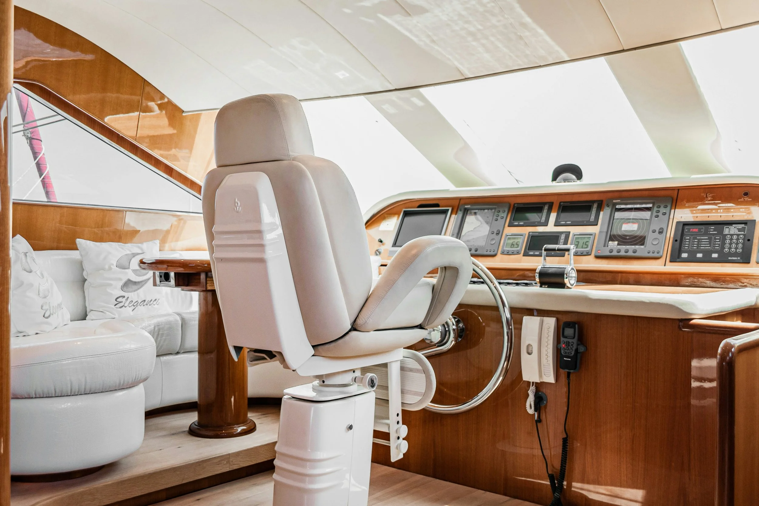 Interior of a yacht cockpit with a white captain's chair, a wooden dashboard with navigation equipment, and a white sofa with decorative pillows.