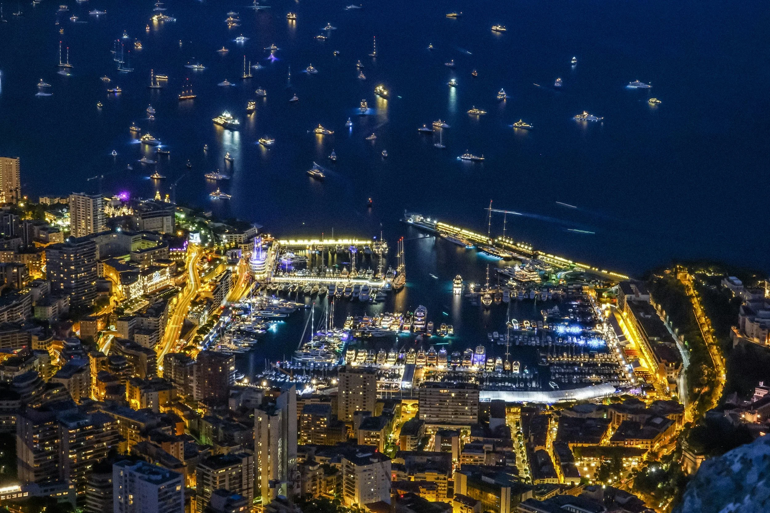 Nighttime aerial view of a bustling city harbor with illuminated buildings, boats, and yachts docked at the marina, with a large body of water filled with numerous boats and ships in the background.