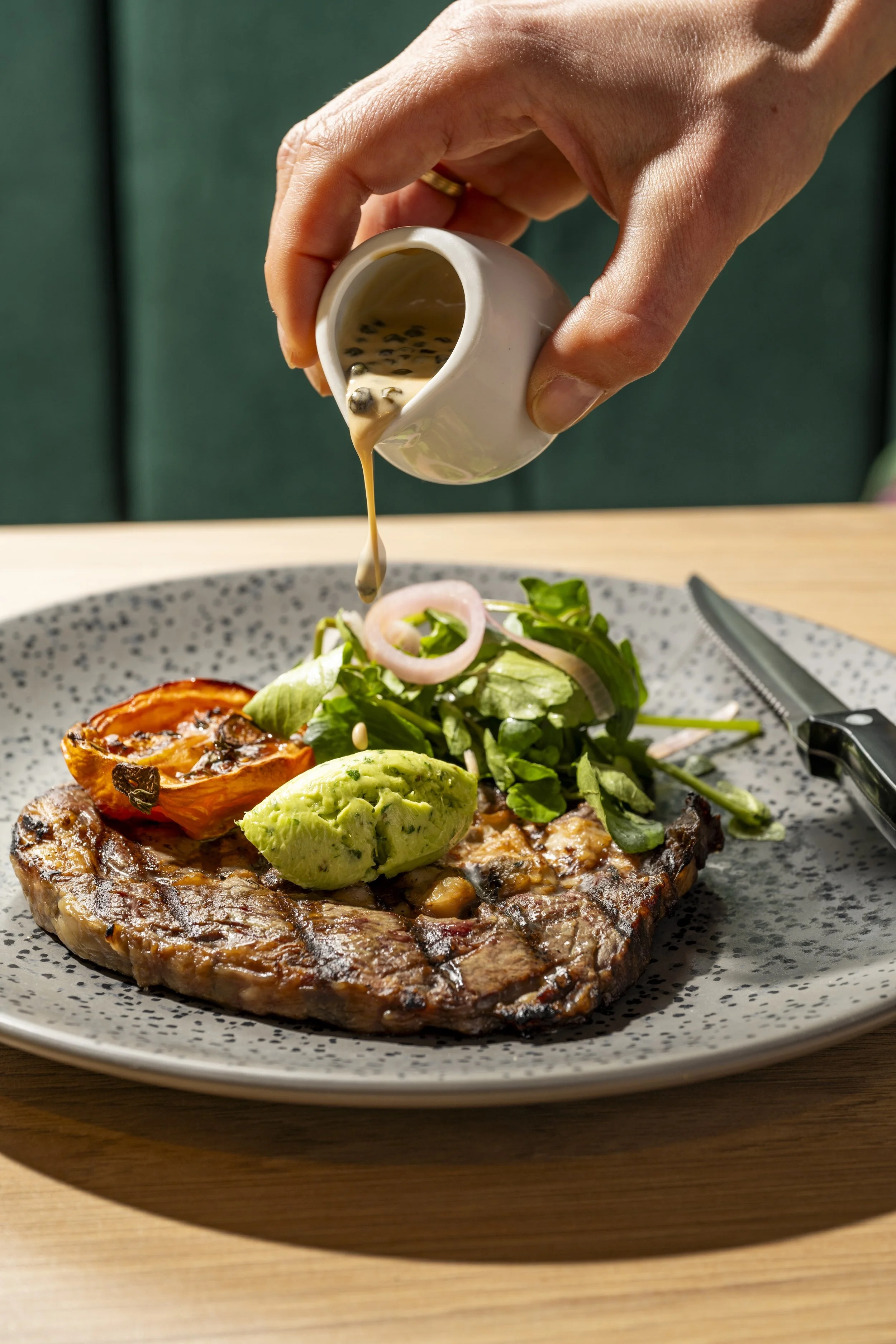 A grilled steak topped with a scoop of guacamole, a grilled tomato, and a vegetable salad with greens, onions, and cucumbers, with a hand pouring, or about to pour, salad dressing on top.