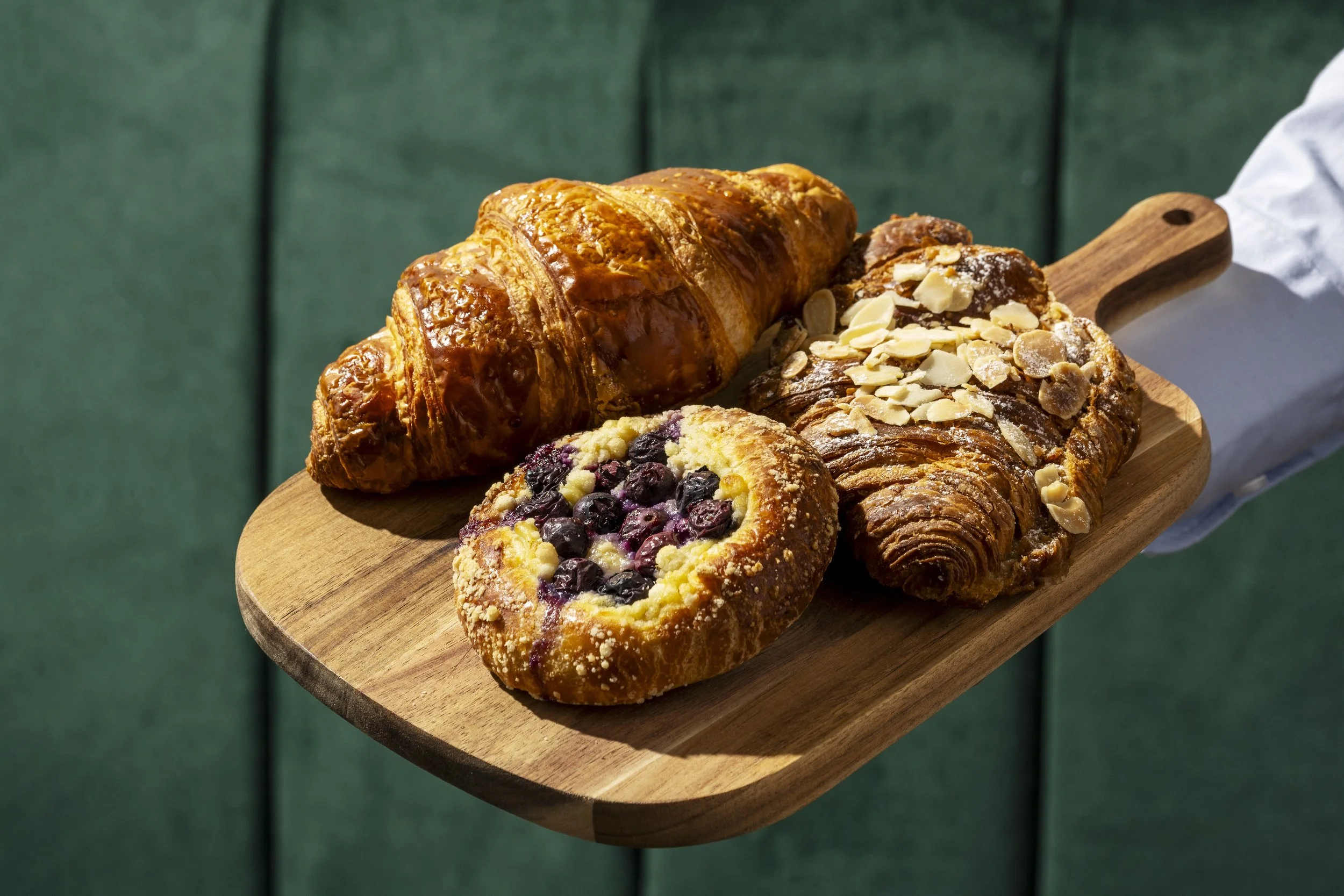 Assorted baked goods on a wooden serving board, including a croissant, fruit Danish pastry with blueberries, almond-topped pastry, and other pastries, with a green cushioned background.