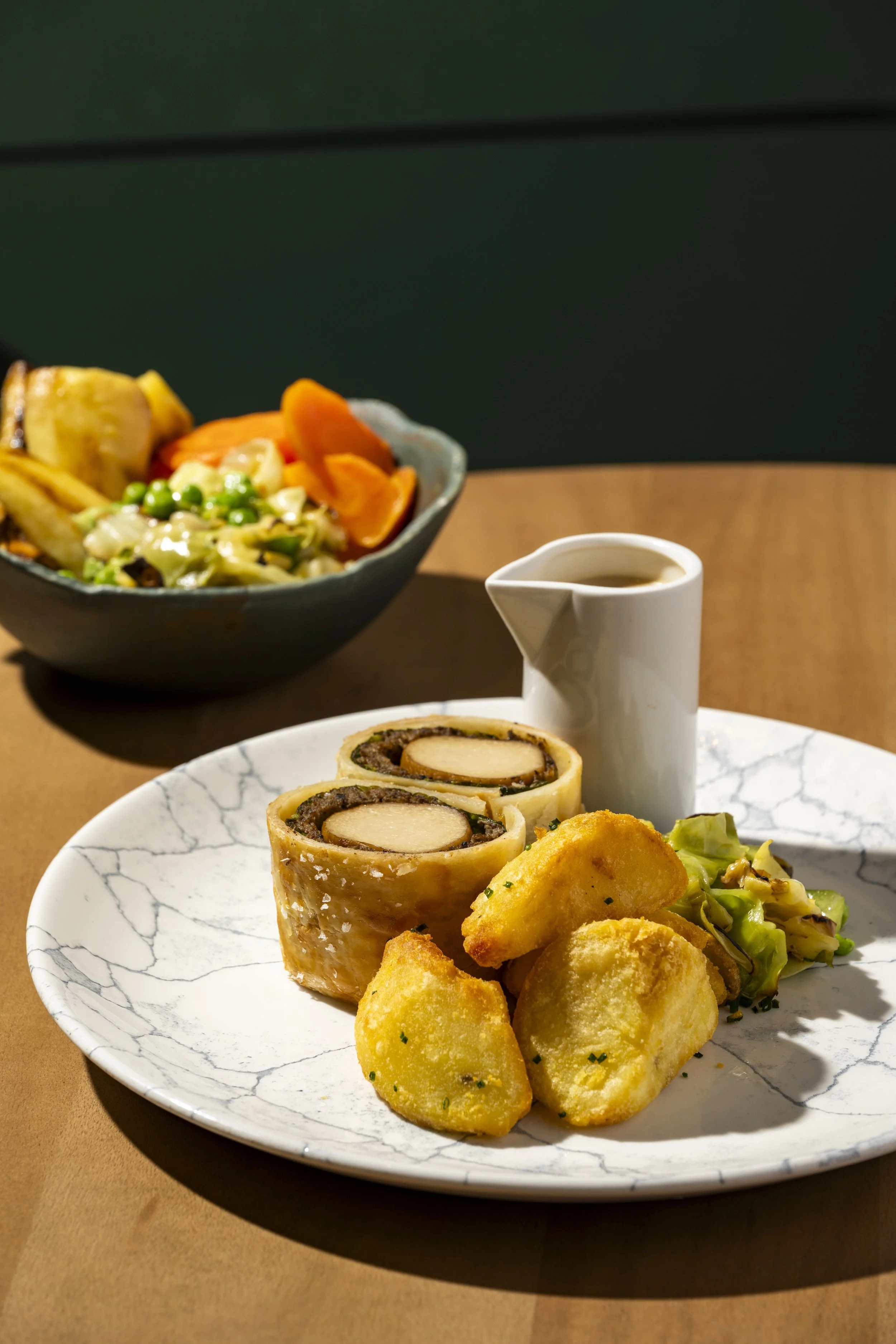 Close-up of a white marble-patterned plate with potato croquettes, a vegetable salad, and a rolled beef dish. In the background, a bowl of mixed vegetables and a small white pitcher.