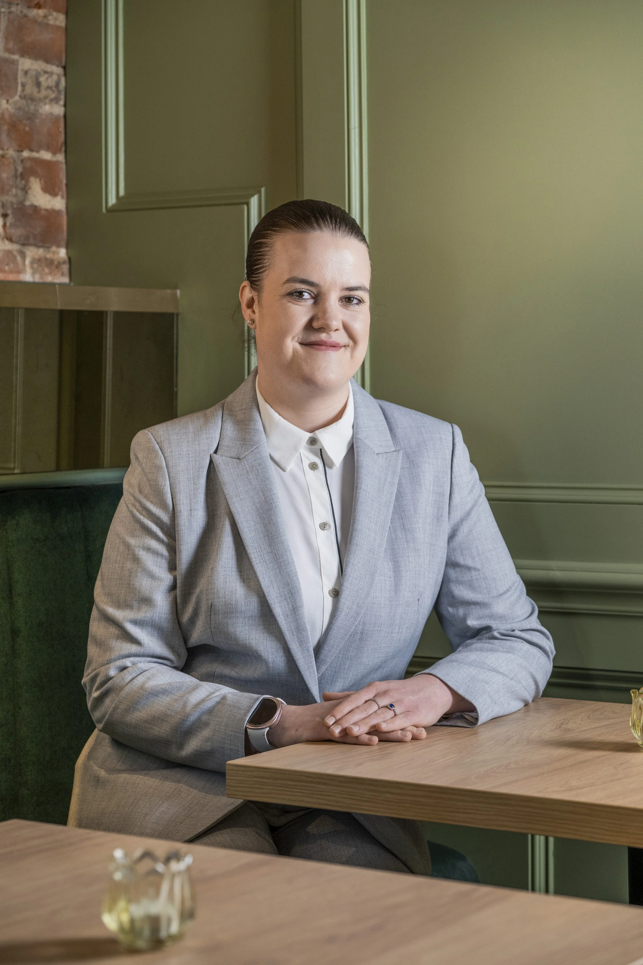 Zuzanna  in a light gray suit sitting at  a table in the main dining room.  She has her hands folded on the table and is smiling at the camera.