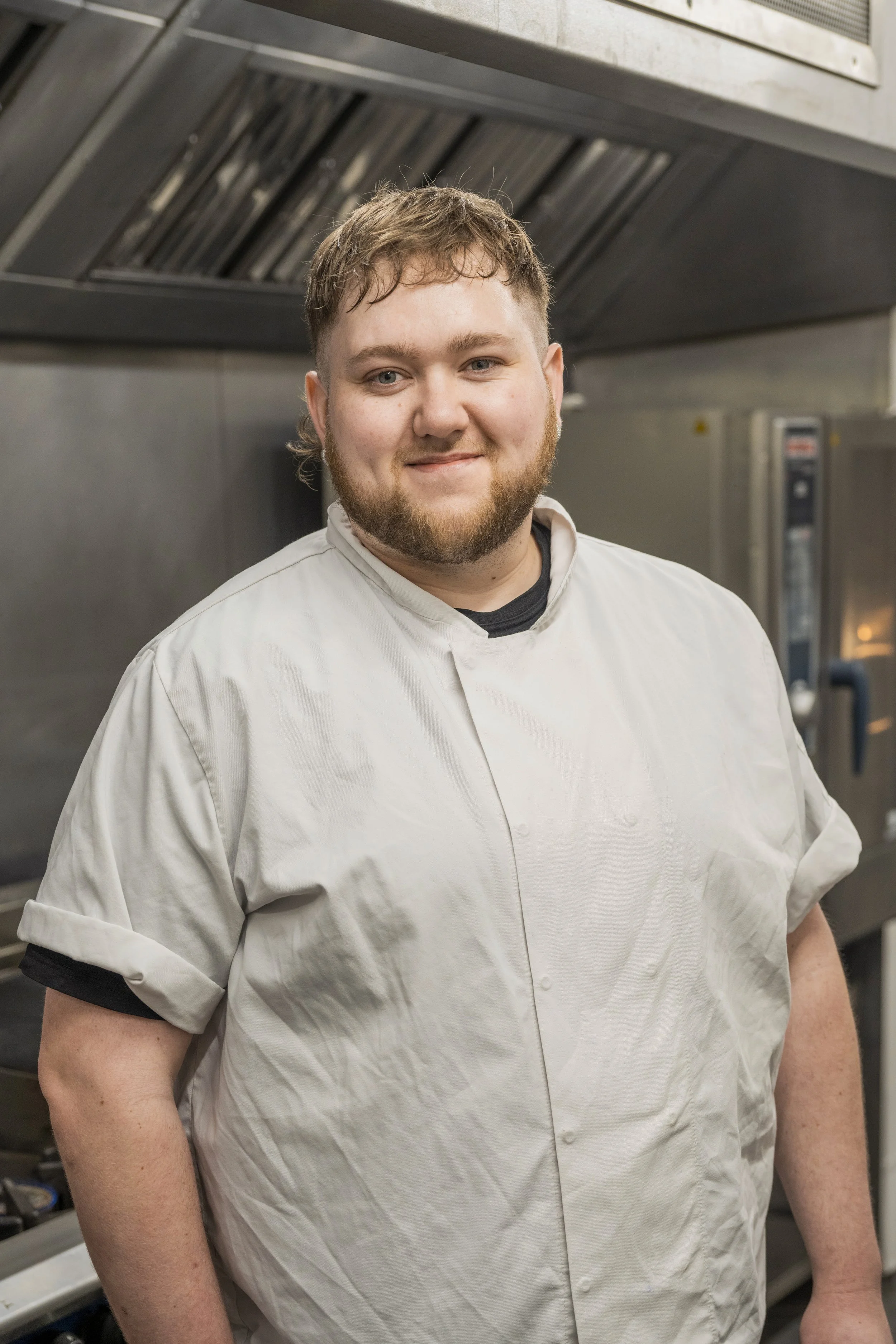 A smiling chef with short brown hair and a beard wearing a white chef's jacket in a commercial kitchen.