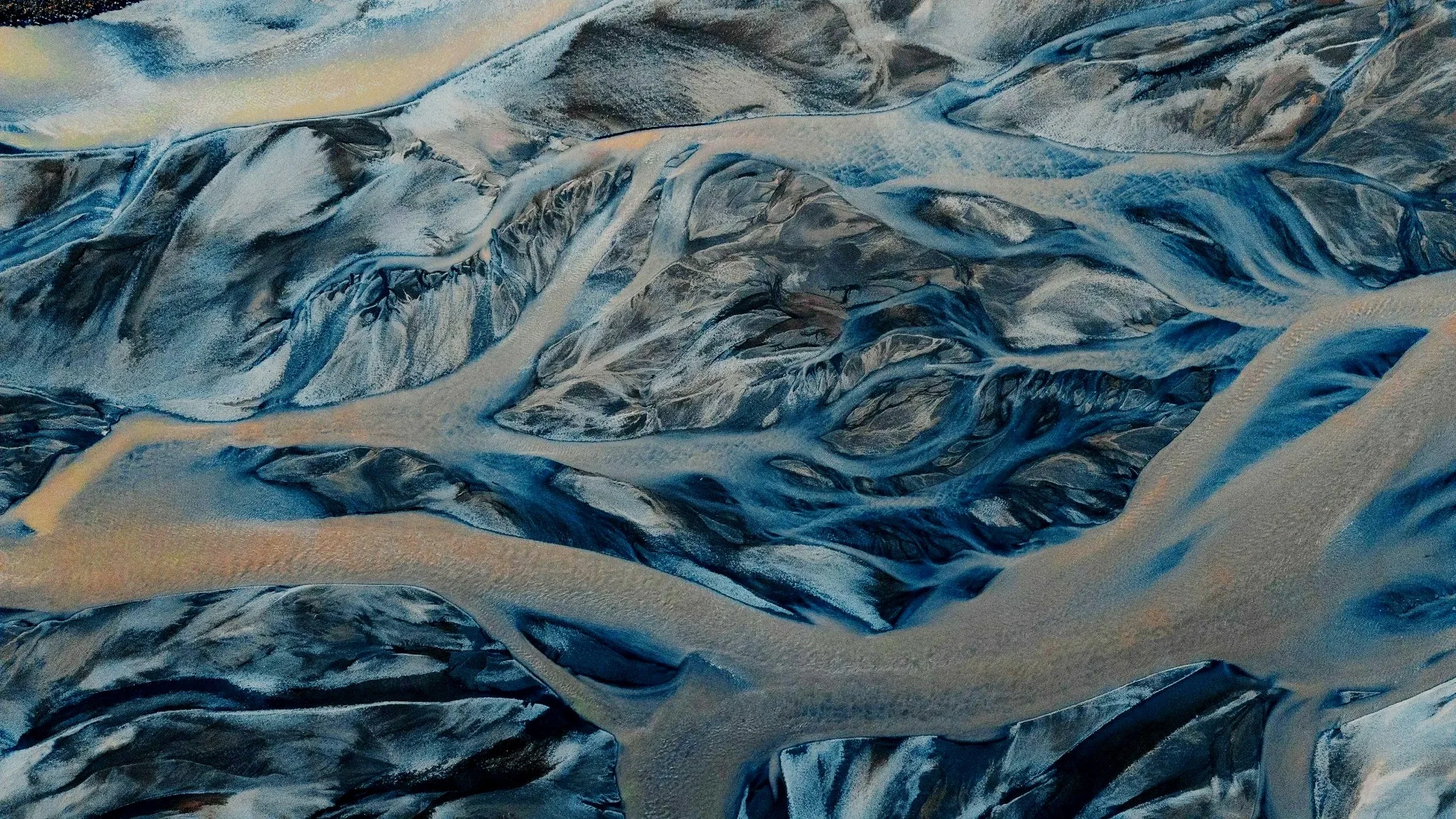 Aerial view of mountains and glaciers with blue, gray, and beige tones characterized by rugged terrain and deep crevices.