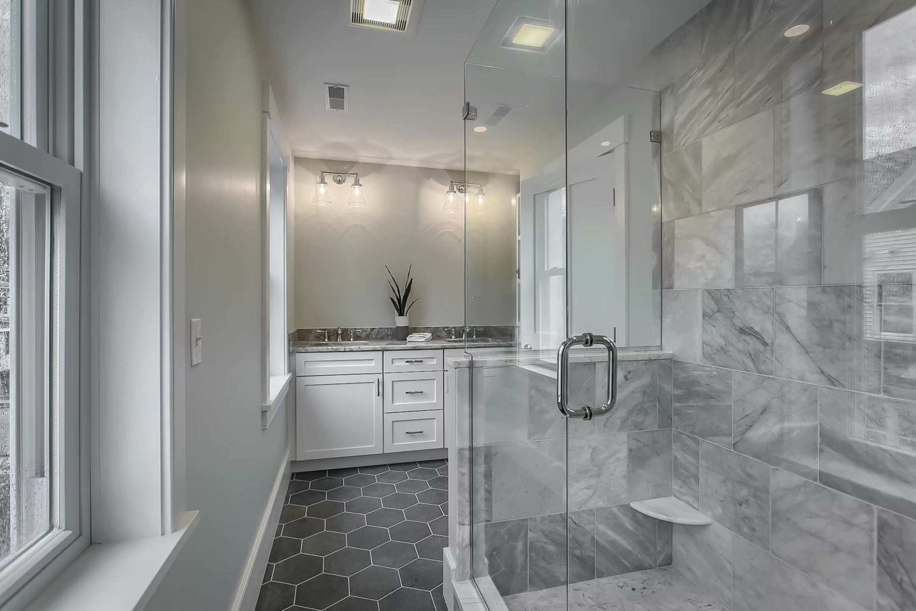 Modern bathroom with a glass shower enclosure, marble tiles on the wall, gray hexagonal floor tiles, a white vanity with a gray marble countertop, and a window allowing natural light.