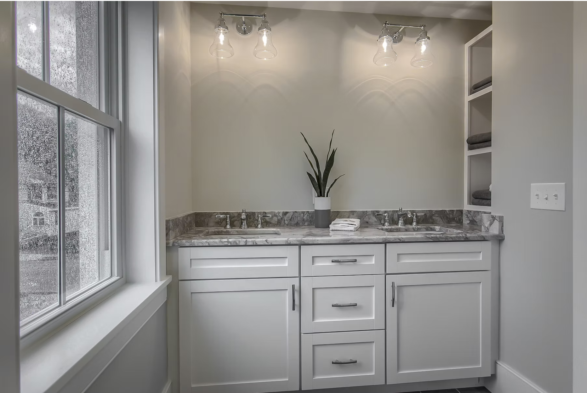 A bathroom vanity with a marble countertop, two sinks, and a tall potted plant in the center. There are folded towels on the counter, open shelving with towels on the right, and a window on the left with raindrops on the glass. Overhead, there's a li