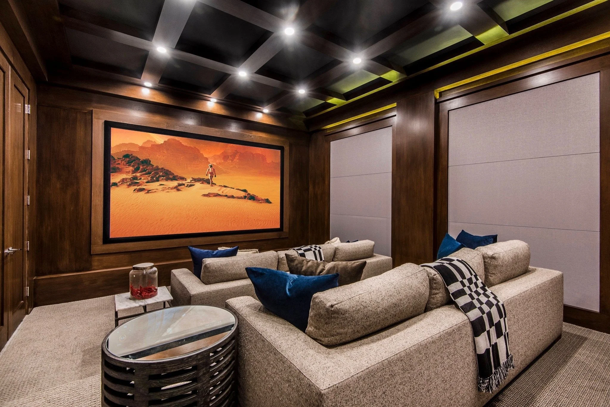 Luxury home theater room with large framed desert landscape painting on wood-paneled wall, beige sectional sofa with blue and brown pillows, black and white checkered throw blanket, round glass-top side table, and recessed ceiling lights.