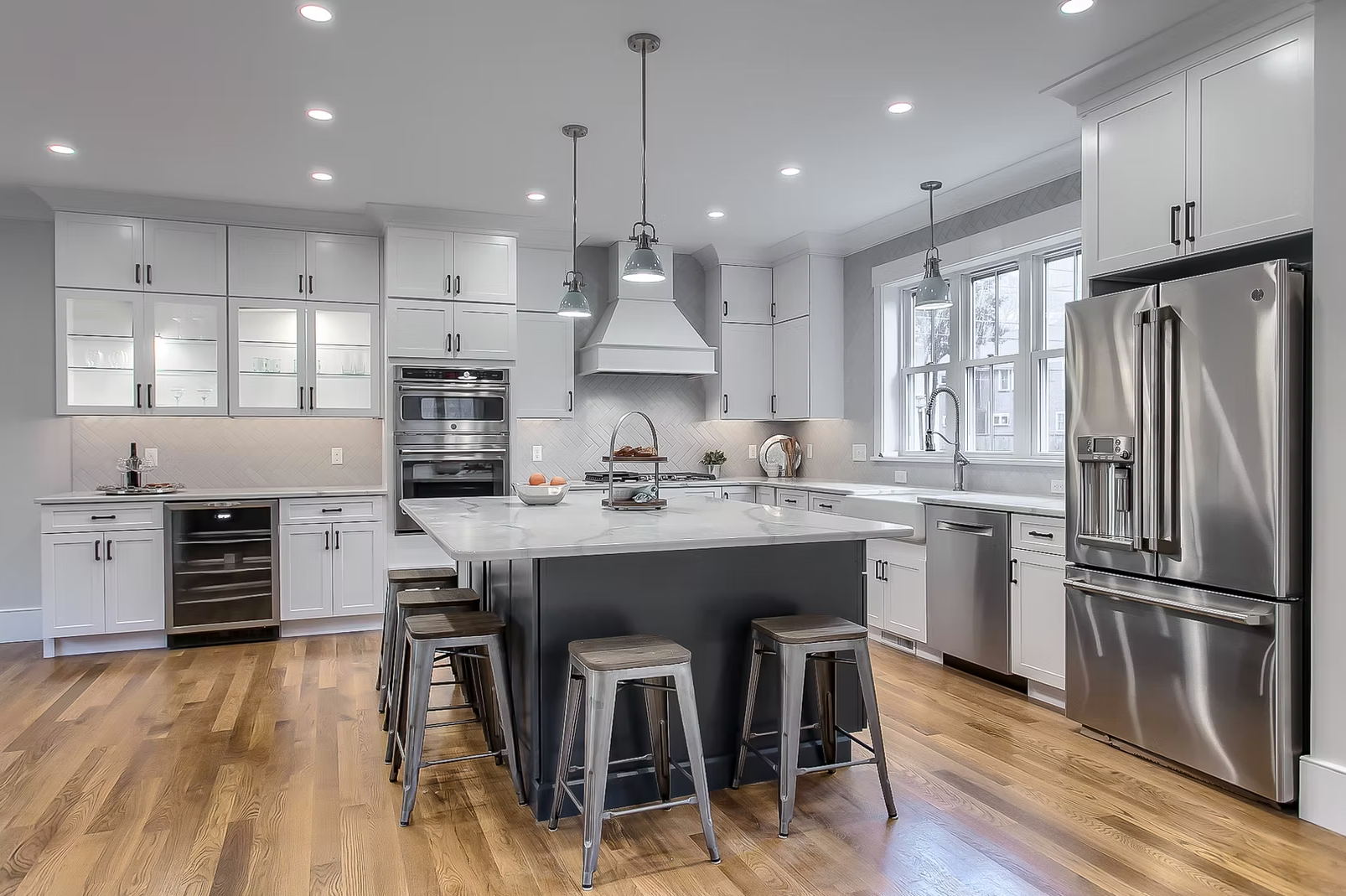 Modern kitchen with white cabinets, stainless steel appliances, a central island with stools, hardwood floors, and large windows providing natural light.