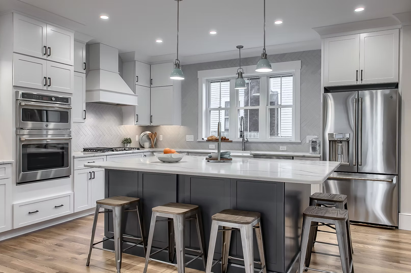 Modern kitchen with white cabinetry, stainless steel appliances, a large central island, and hardwood floors. There are three pendant lights hanging above the island, and a window behind the sink.
