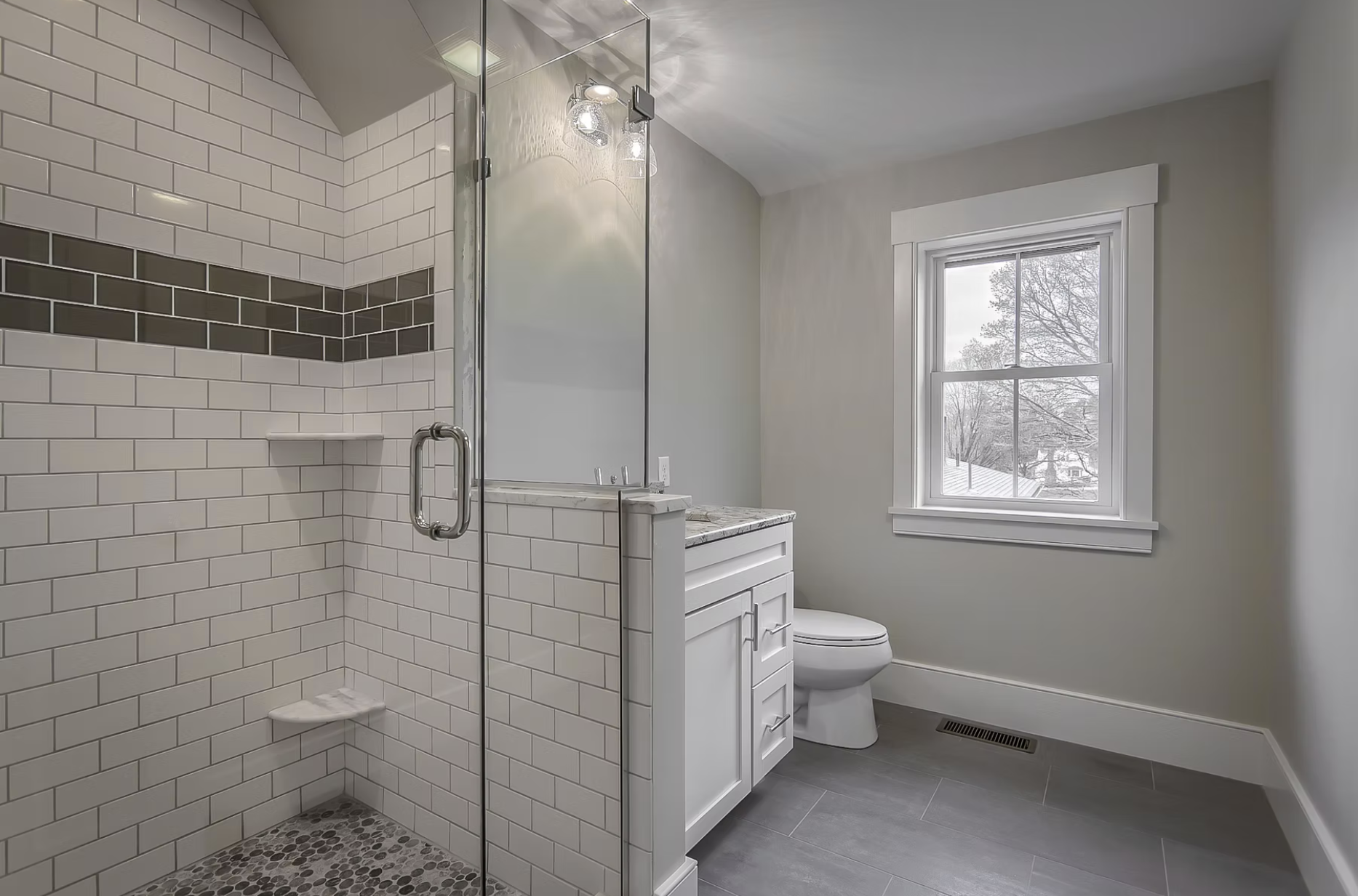 A modern bathroom featuring a glass-enclosed shower with white subway tiles, a gray accent stripe, and pebble floor, a white vanity with marble countertop, a toilet, and a window with a winter scene outside.